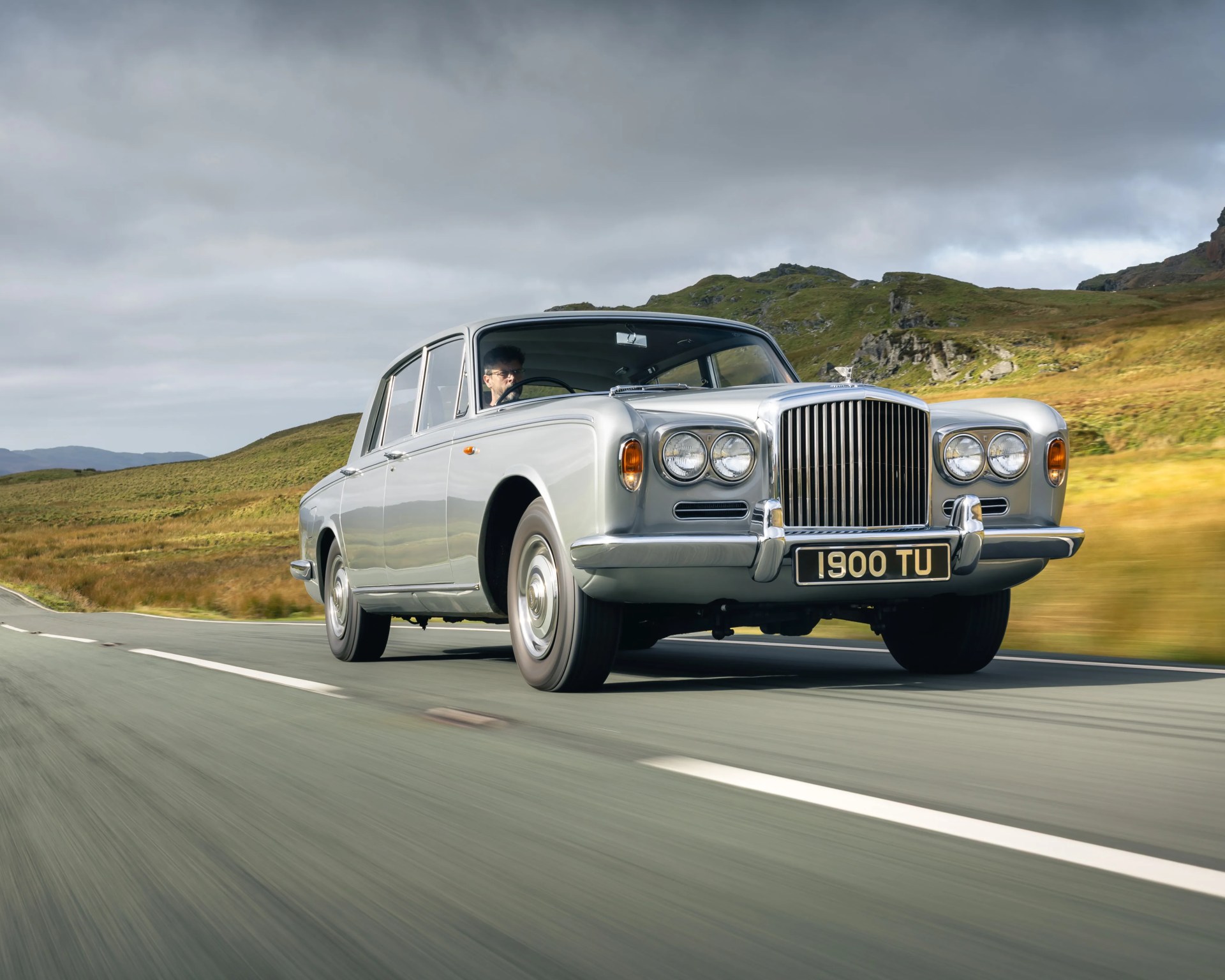 Silver classic luxury sedan driving on a rural road with grassy hills and cloudy sky in the background.