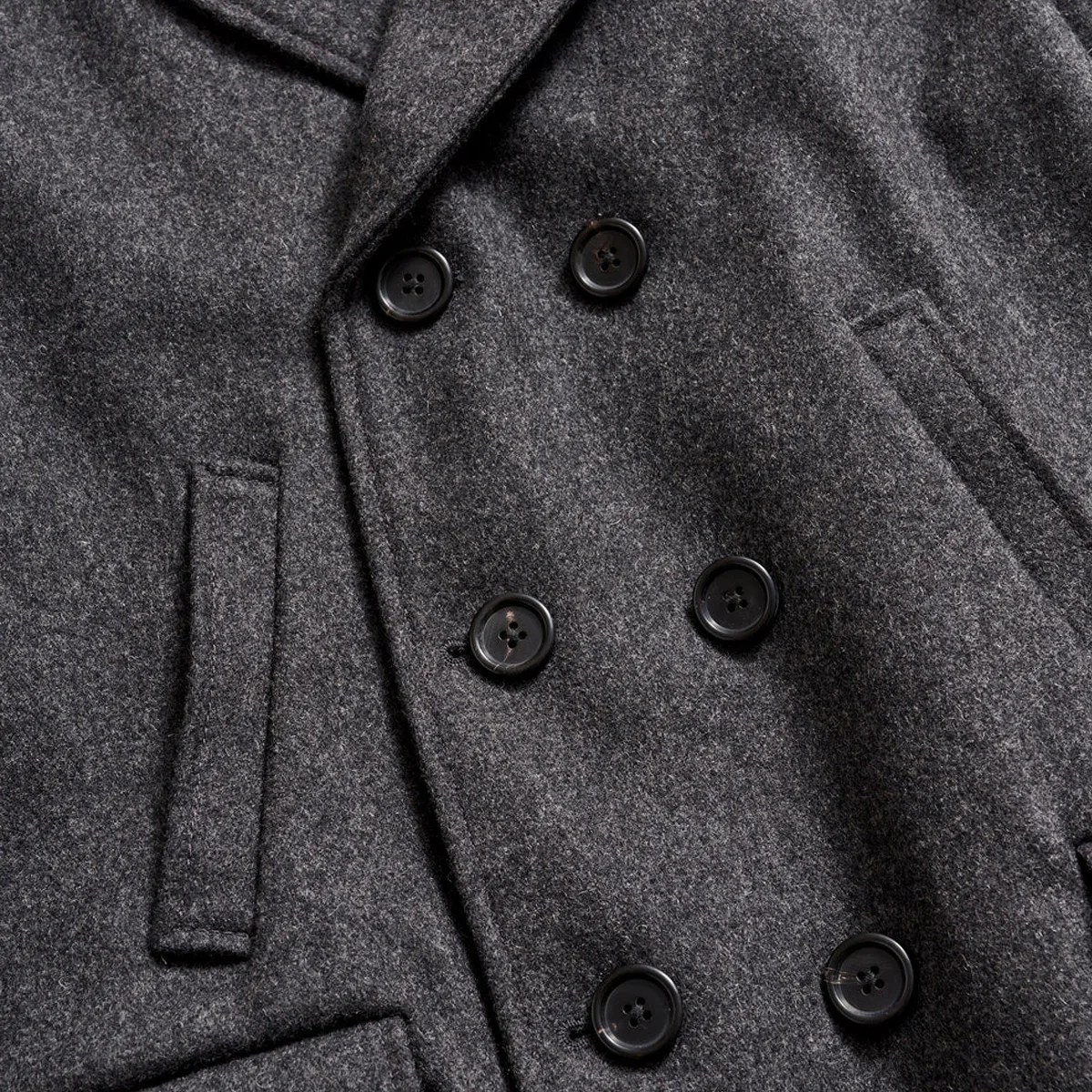 Close-up of a dark gray wool coat with six black buttons and two vertical slit pockets.