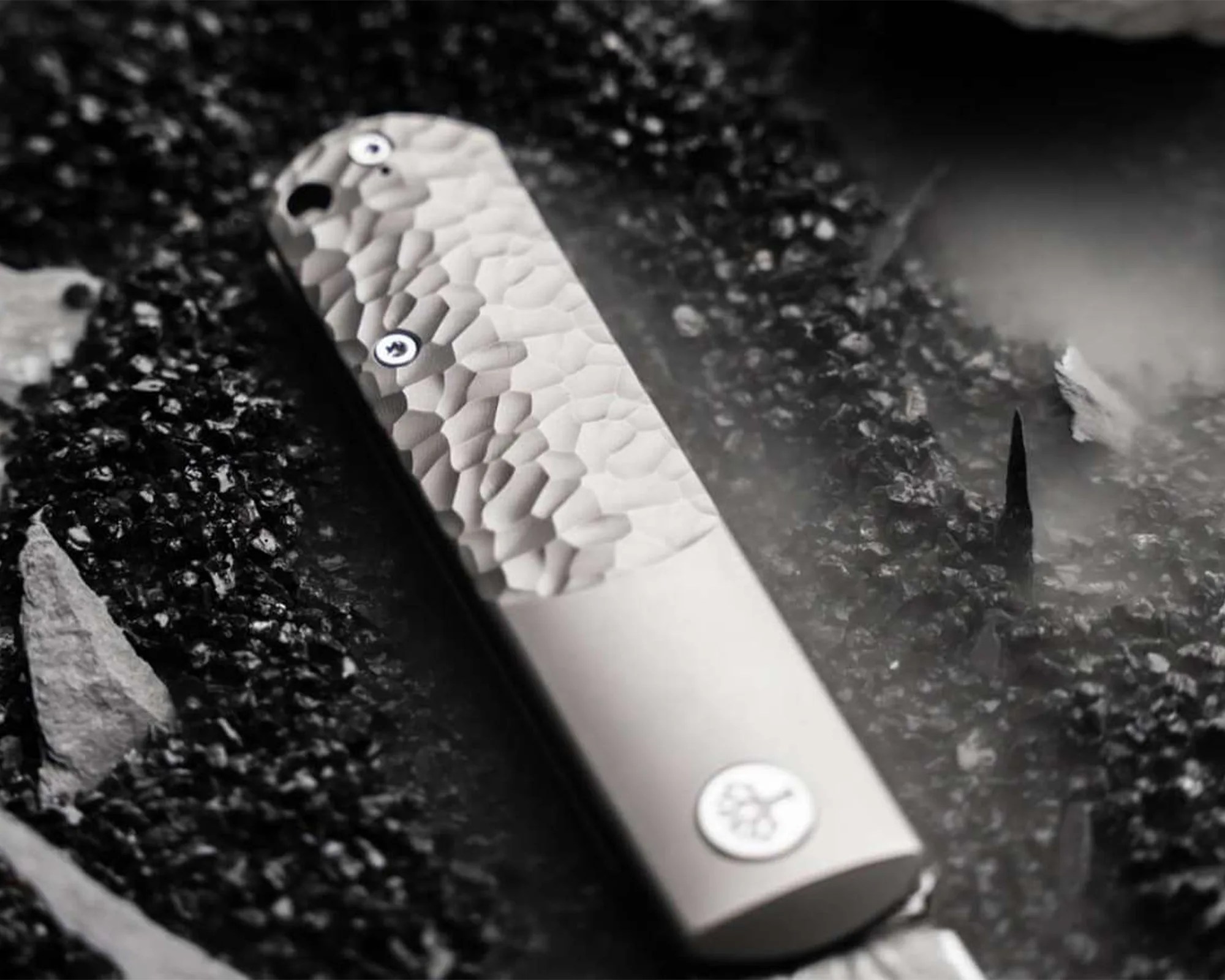 Silver metal pocket knife with a textured hammered finish on the handle, lying on dark gravel.