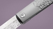 Close-up of a silver folding knife with a textured handle and an engraved blade featuring a tree logo on the handle.