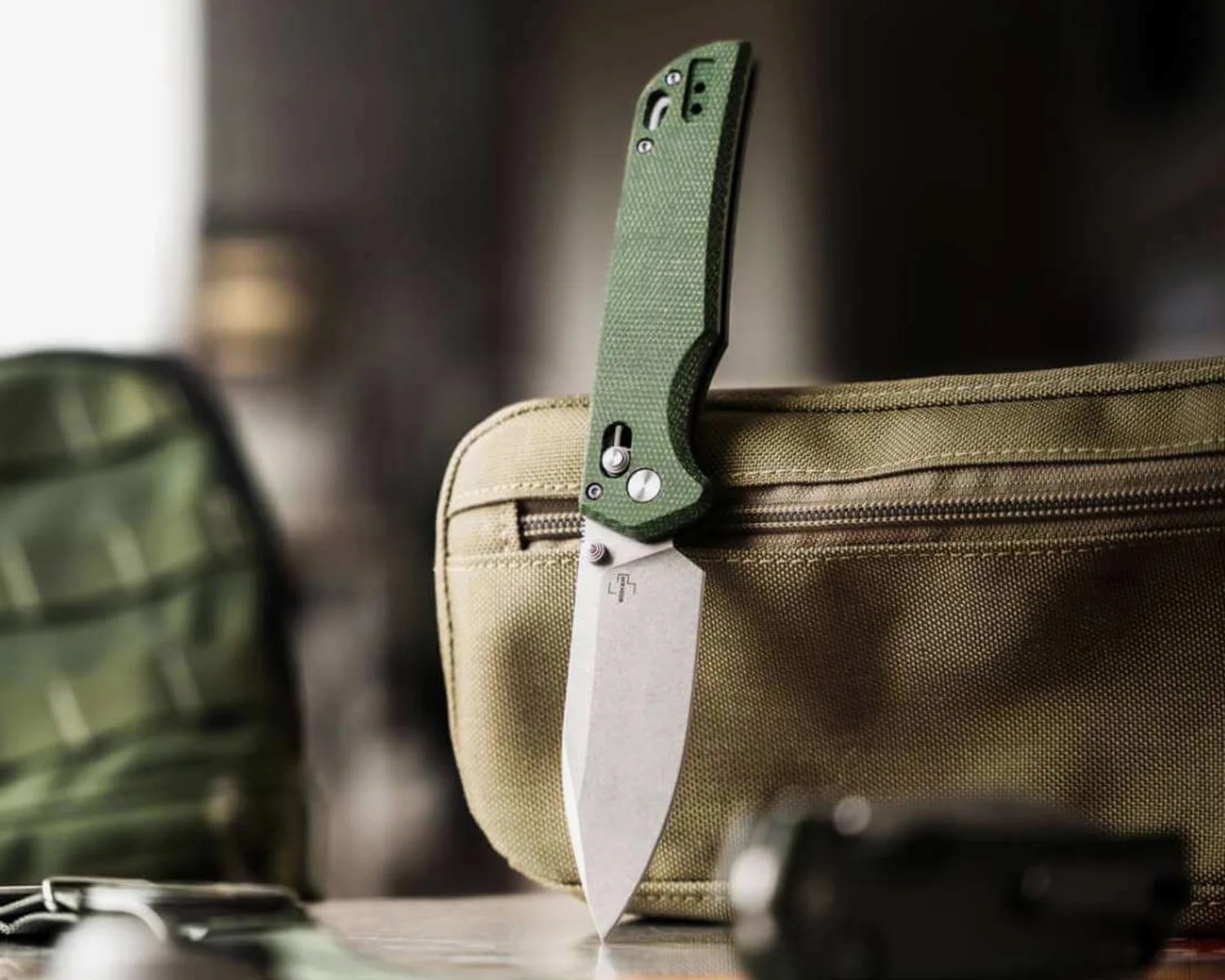 Folding knife with a green textured handle and a stonewashed blade standing upright on a tan fabric pouch.