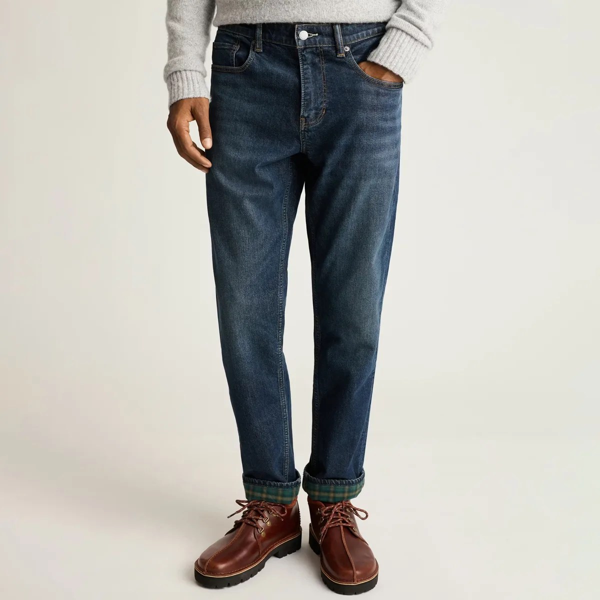 Dark blue denim jeans with rolled cuffs showing green plaid lining, worn with brown leather boots and a gray sweater.