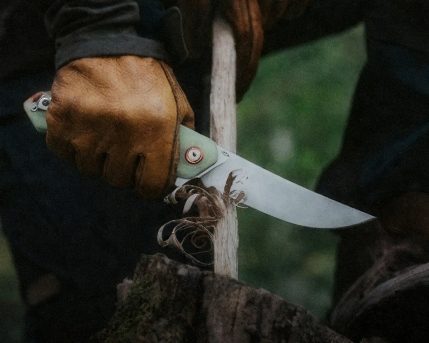 Person wearing brown leather gloves carving a wooden stick with a fixed-blade knife outdoors.