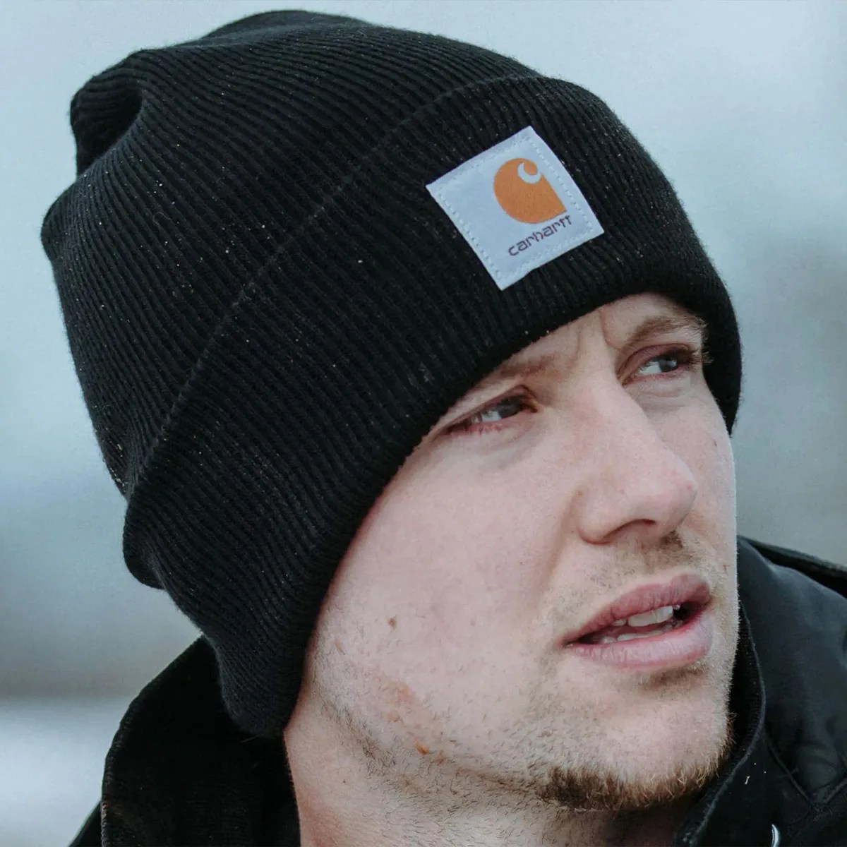 Black ribbed knit beanie with a white and orange Carhartt logo patch worn by a person.