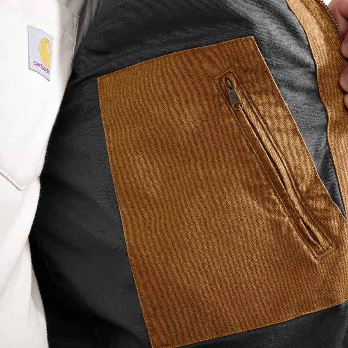Brown fabric inner pocket with zipper on a black jacket lining, worn by a person in a white Carhartt shirt.