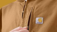 Close-up of a hand zipping the front pocket of a brown Carhartt jacket with a visible logo patch.