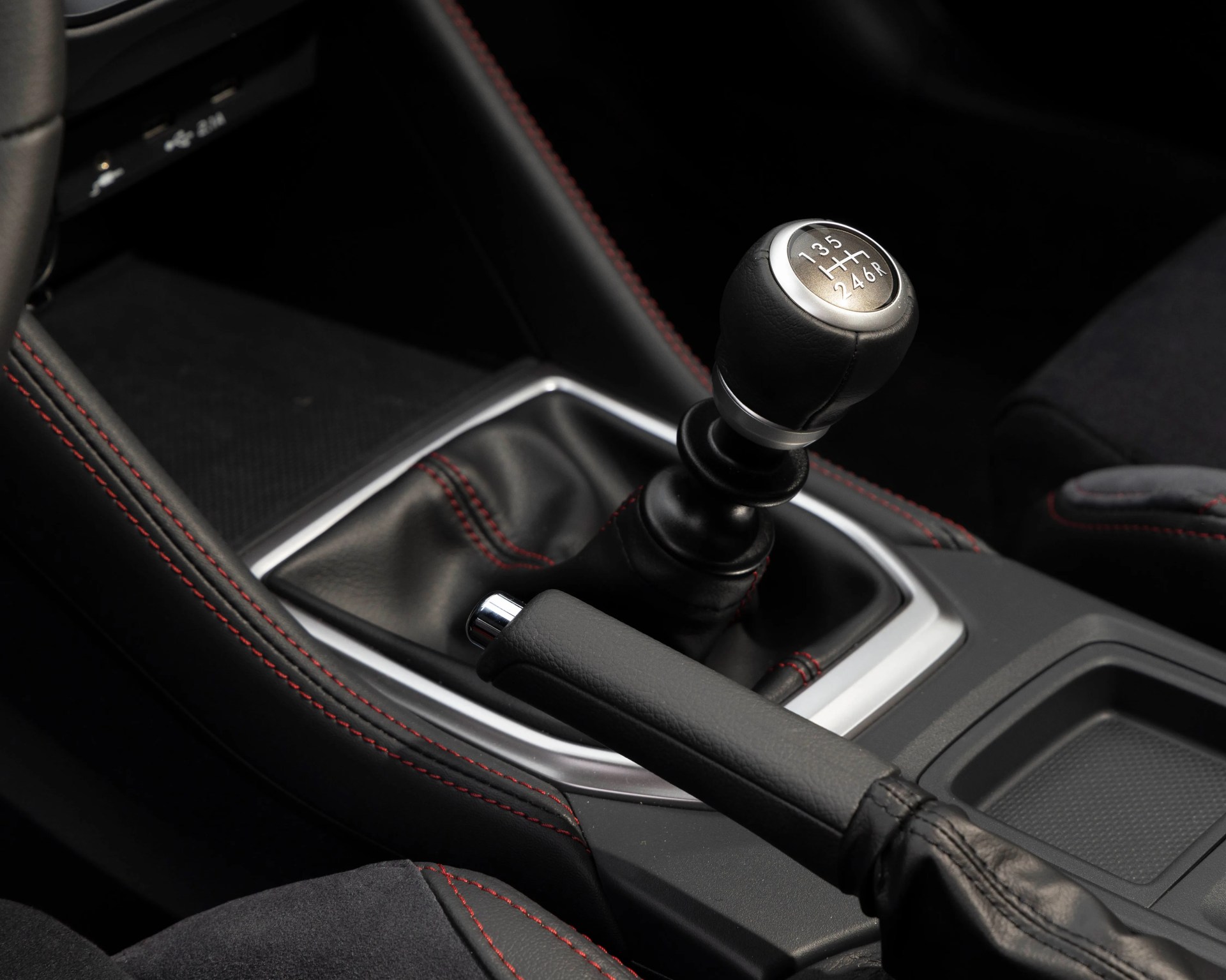 Black leather manual gear shift knob with silver top and red stitching on black leather car interior.
