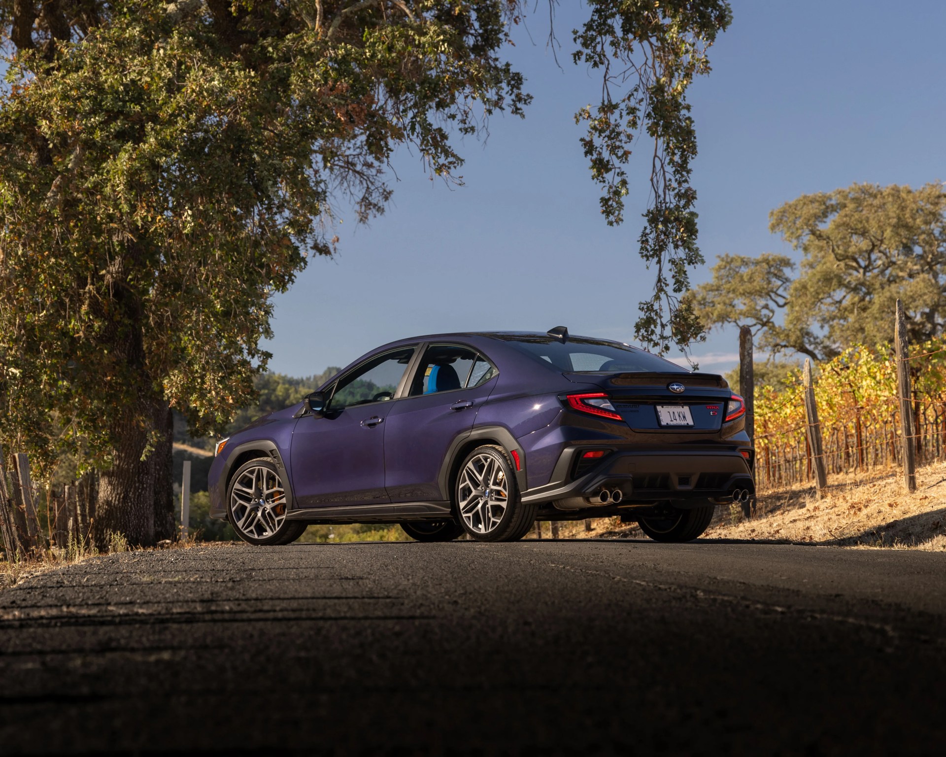 Purple Subaru WRX sedan with quad exhaust pipes parked on a rural road under trees.