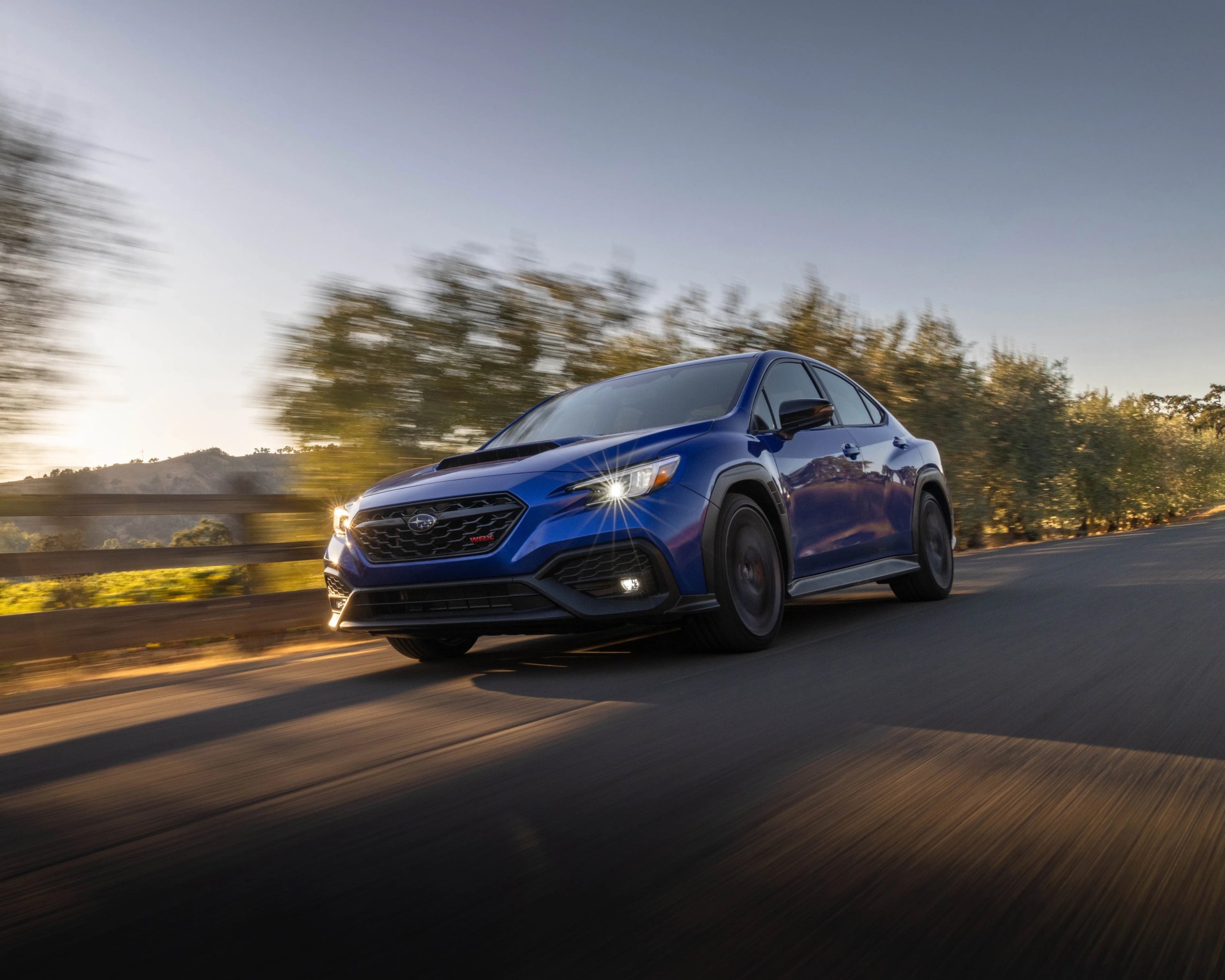 Blue Subaru WRX sedan driving on a sunlit road with blurred trees in the background.