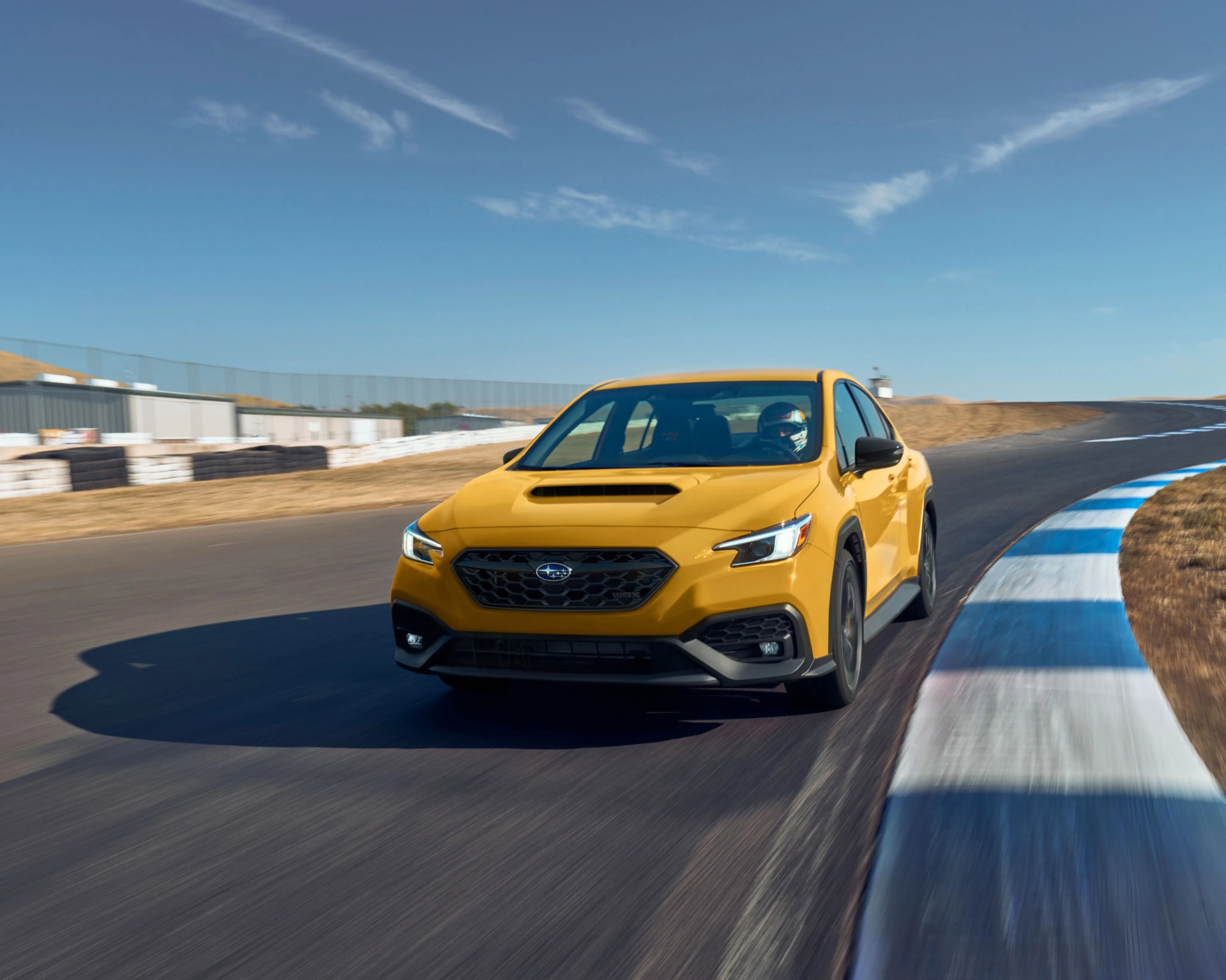 Yellow Subaru WRX driving on a race track with blue and white curbs under a clear sky.
