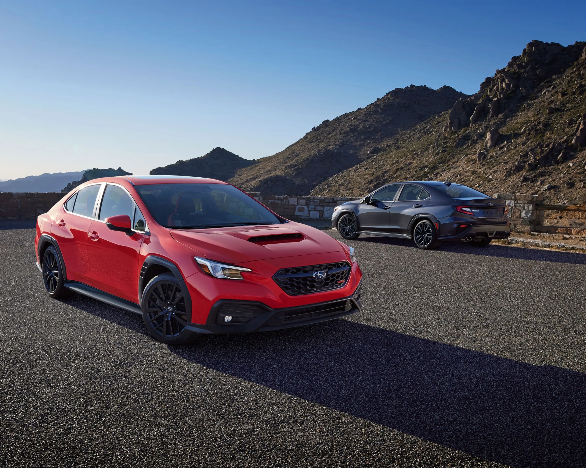 Red Subaru WRX sedan with black wheels parked on asphalt with a gray Subaru WRX sedan in the background near rocky hills.