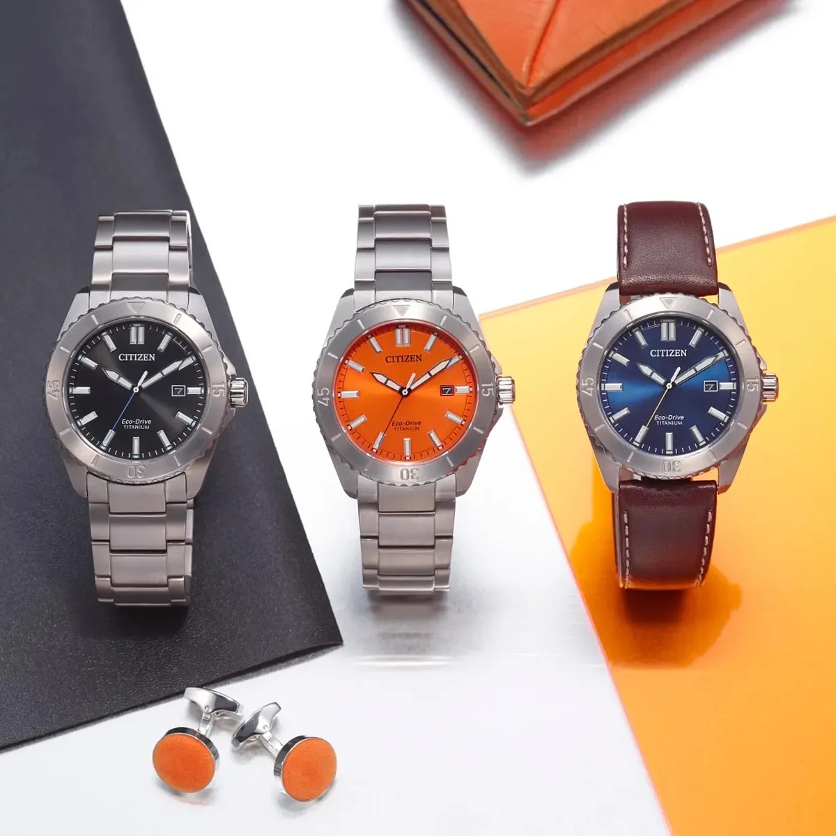 Three Citizen Eco-Drive titanium watches with black, orange, and blue dials, two with metal bands and one with brown leather strap, next to orange cufflinks.