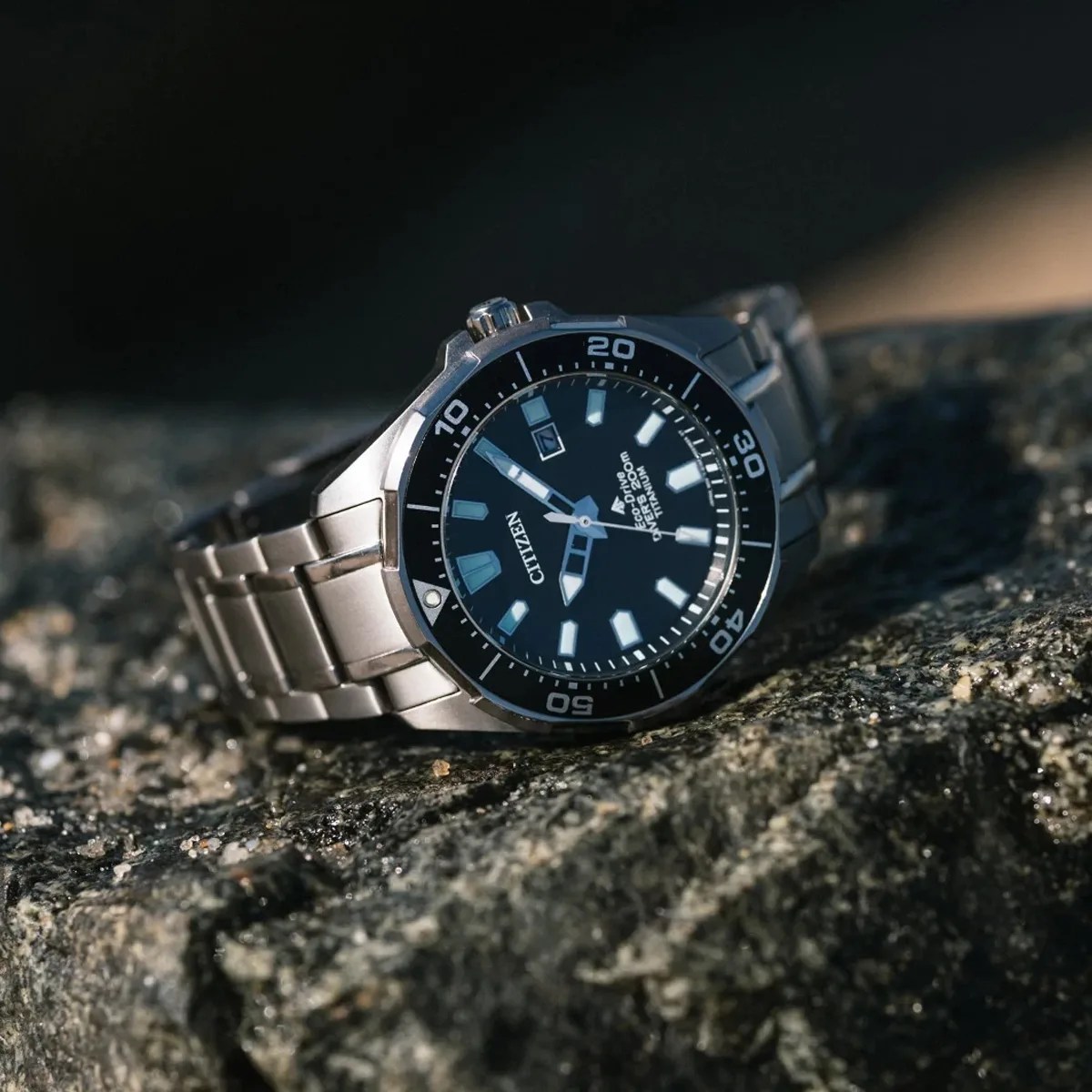 Silver Citizen Eco-Drive titanium wristwatch with black dial and rotating bezel on a rock surface.
