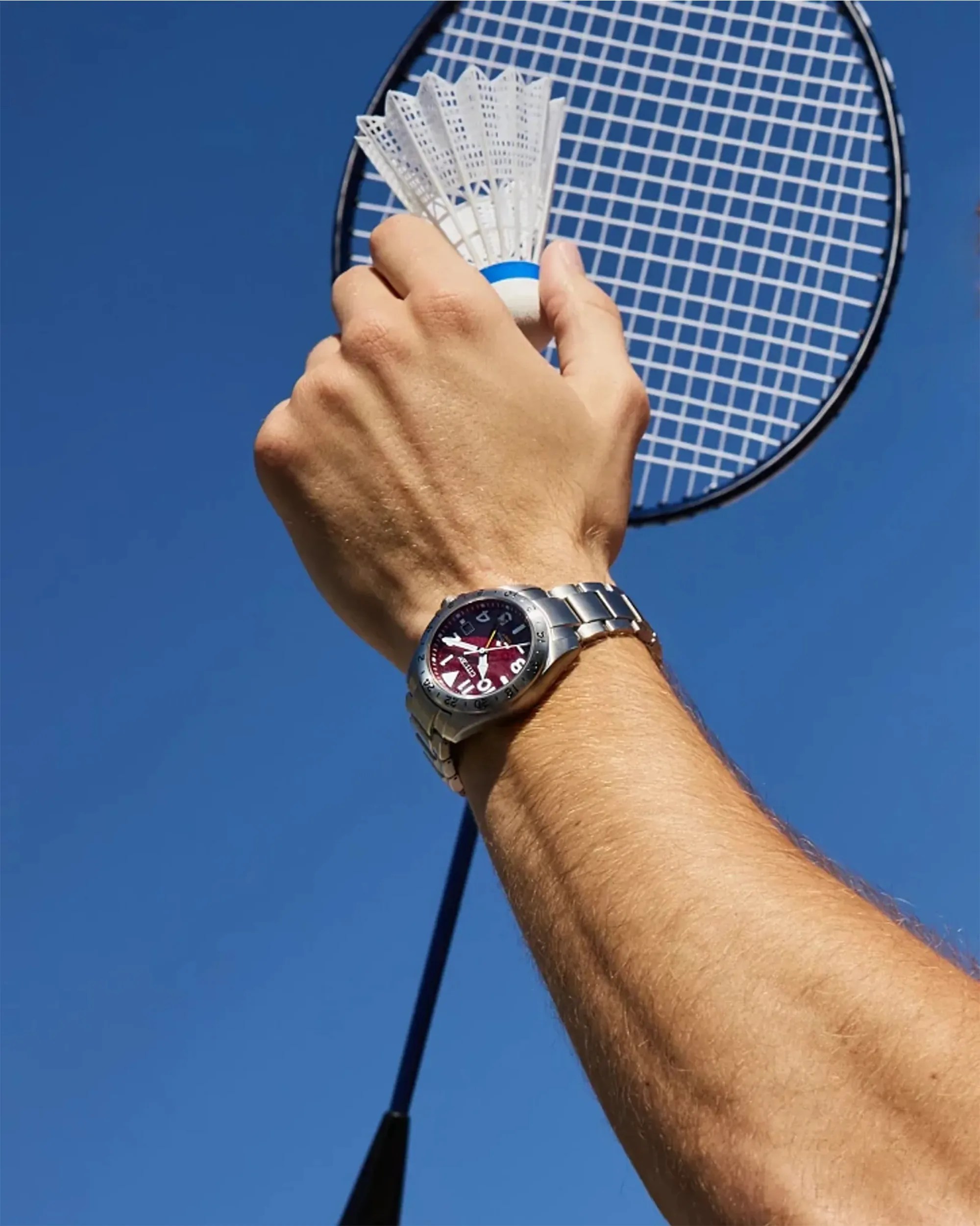 Arm wearing a silver metal wristwatch holding a white badminton shuttlecock and racket against a clear blue sky.