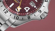 Close-up of a silver metal wristwatch with a textured maroon dial, yellow arrow hand, and date window showing the number 7.
