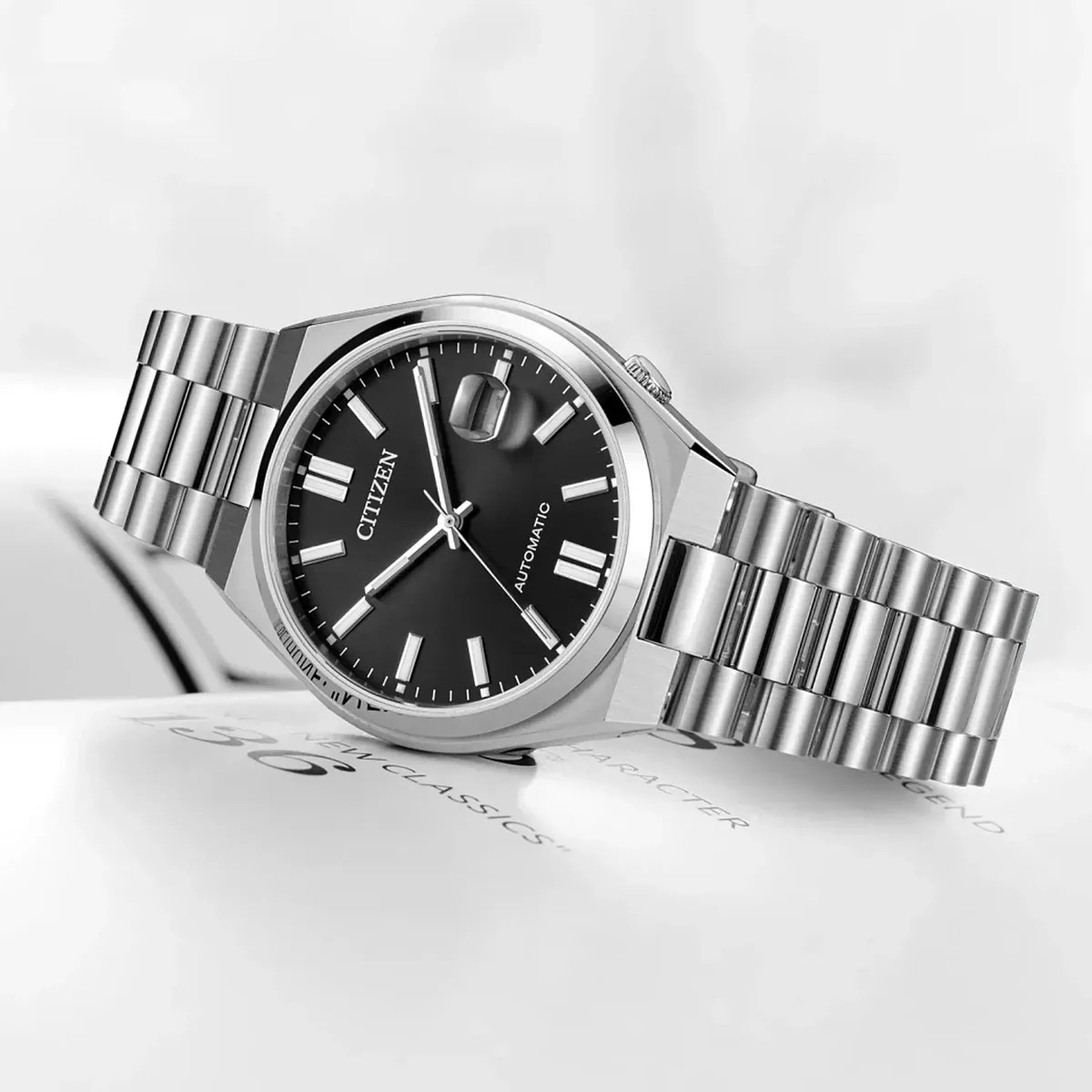 Silver Citizen automatic wristwatch with black dial and metal link bracelet resting on a white surface.