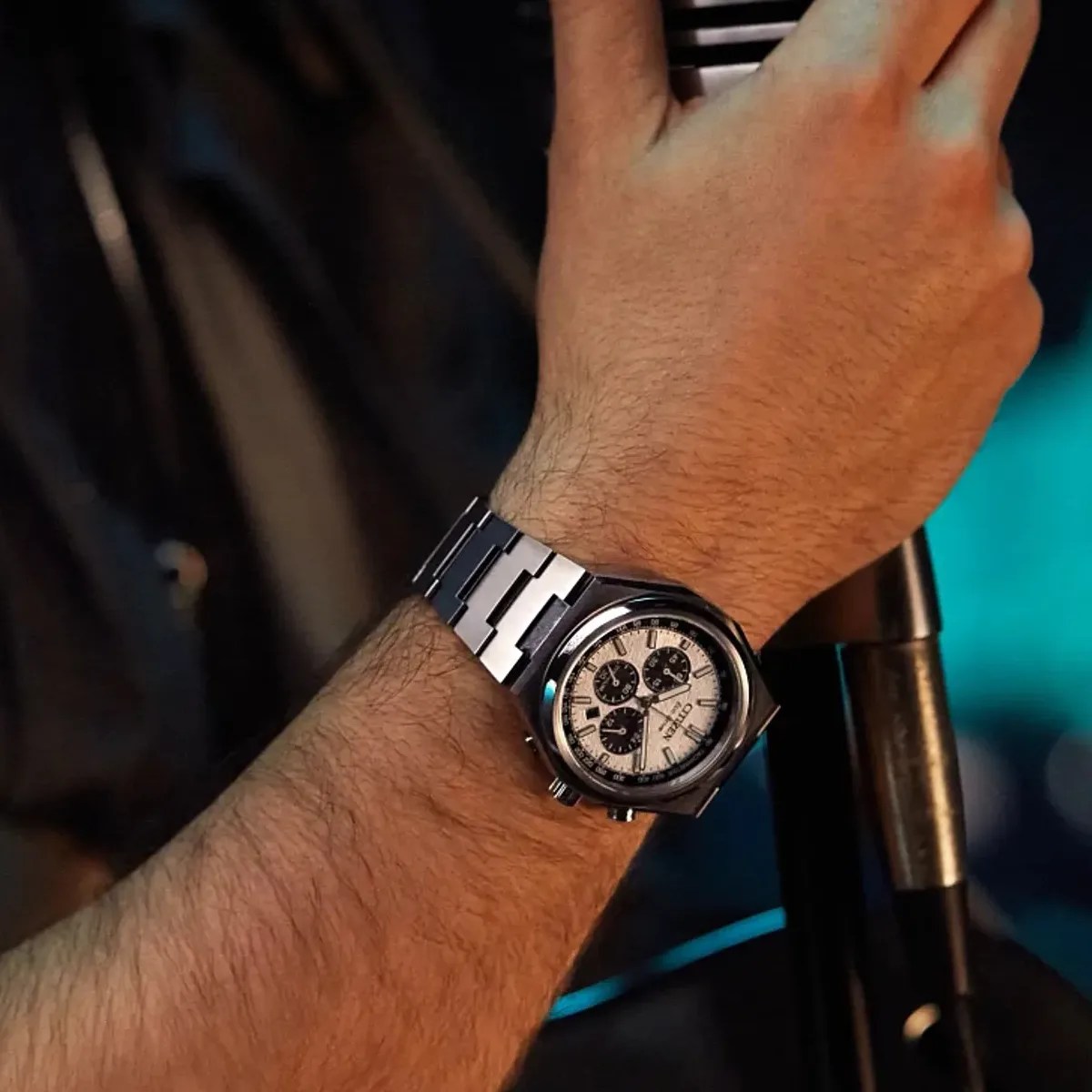 Silver metal wristwatch with a white dial and three black subdials worn on a wrist holding a microphone.