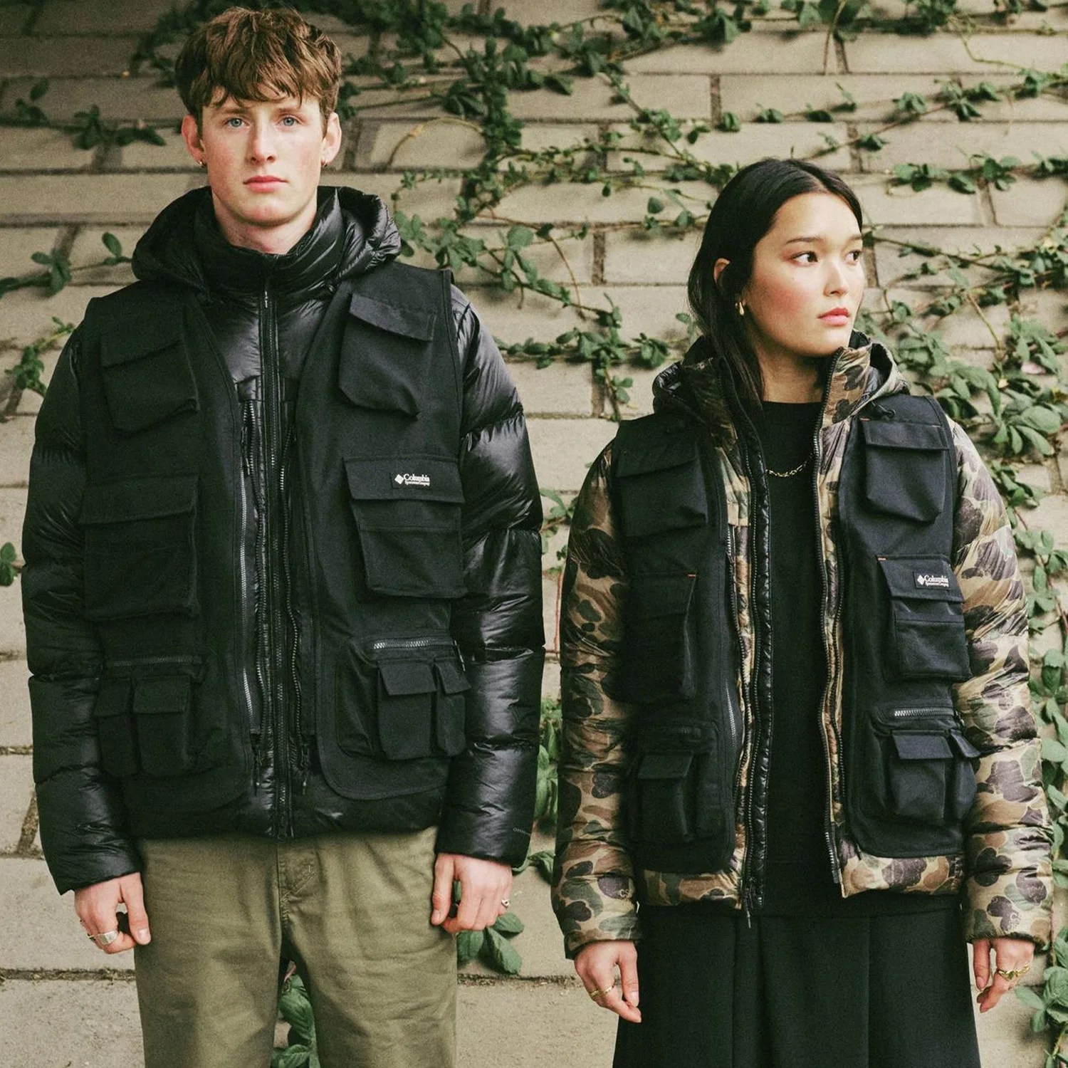 Two people wearing black Columbia utility vests over puffer jackets, one in black and the other in camouflage, standing against a vine-covered stone wall.