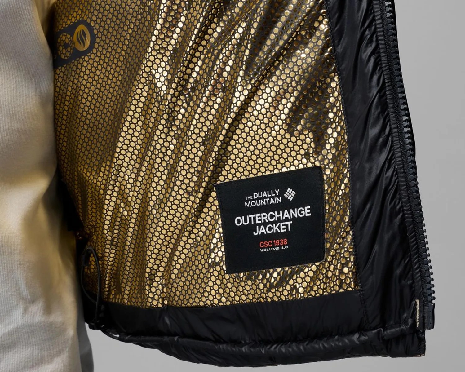 Close-up of a jacket interior with a gold honeycomb-patterned lining and a black label reading "The Dually Mountain Outerchange Jacket CSC 1938 Volume 1.0.