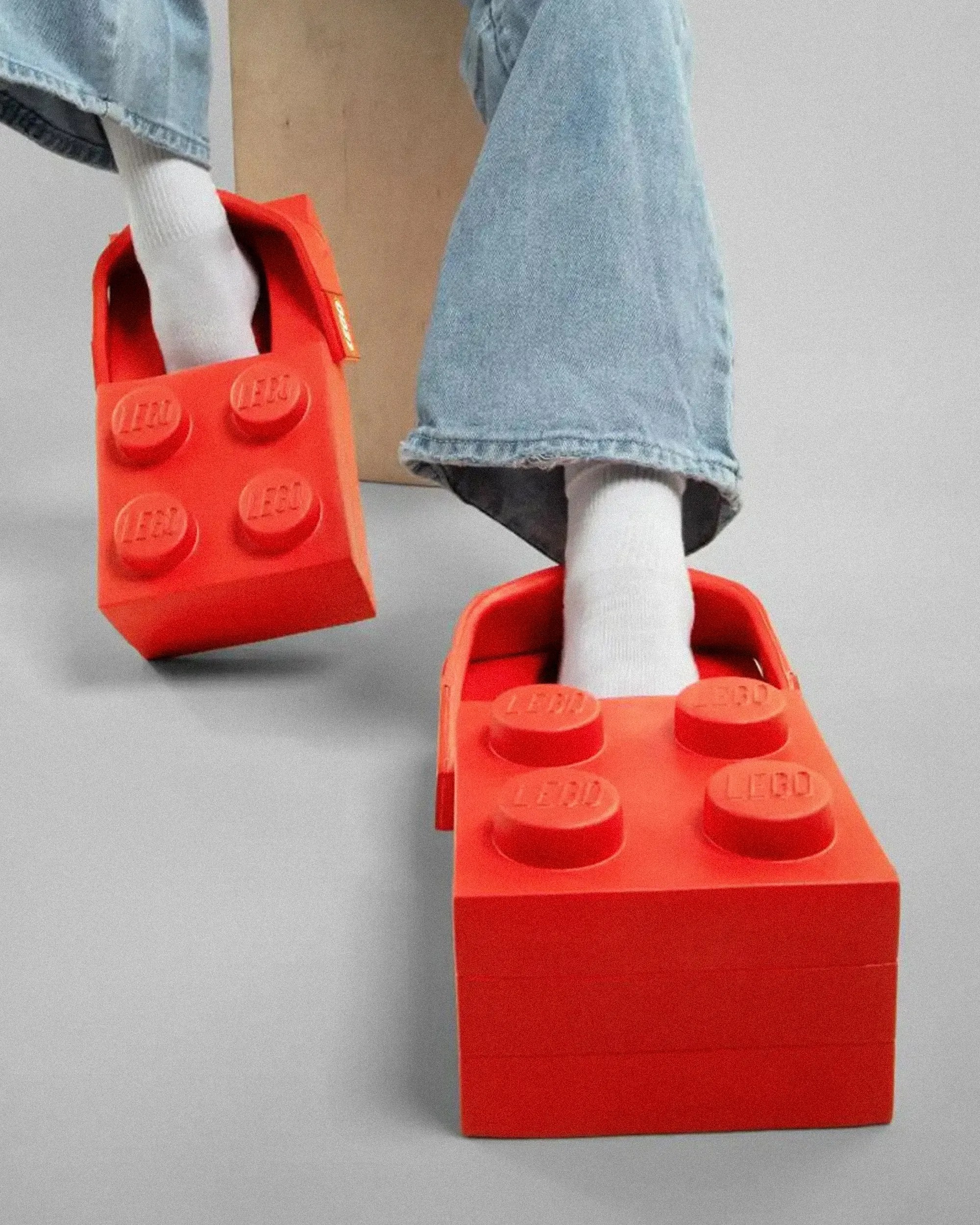 Bright red oversized LEGO brick slippers worn with white socks and light blue jeans.
