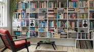 Wall-mounted white bookshelf filled with books, two white speakers, decorative items, and a lower shelf with vinyl records, next to a red cushioned wooden armchair.