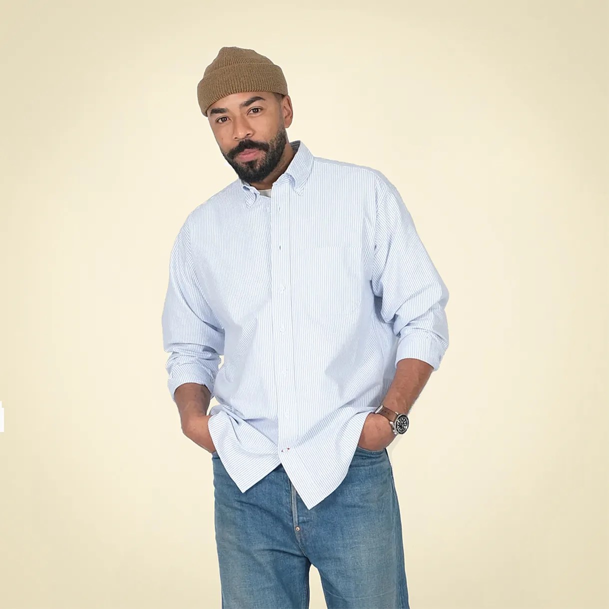 Man wearing a light blue and white striped button-up shirt, blue jeans, brown knit beanie, and wristwatch.