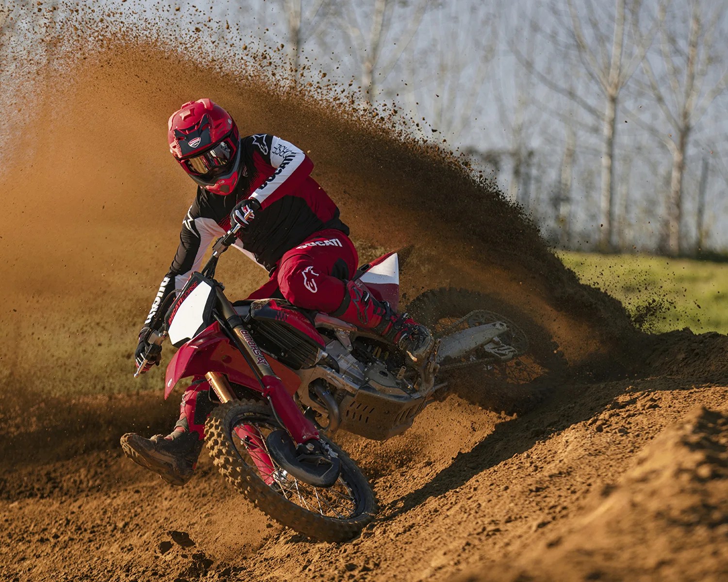 Motocross rider in red gear kicking up dirt while turning on a red dirt bike on a track.