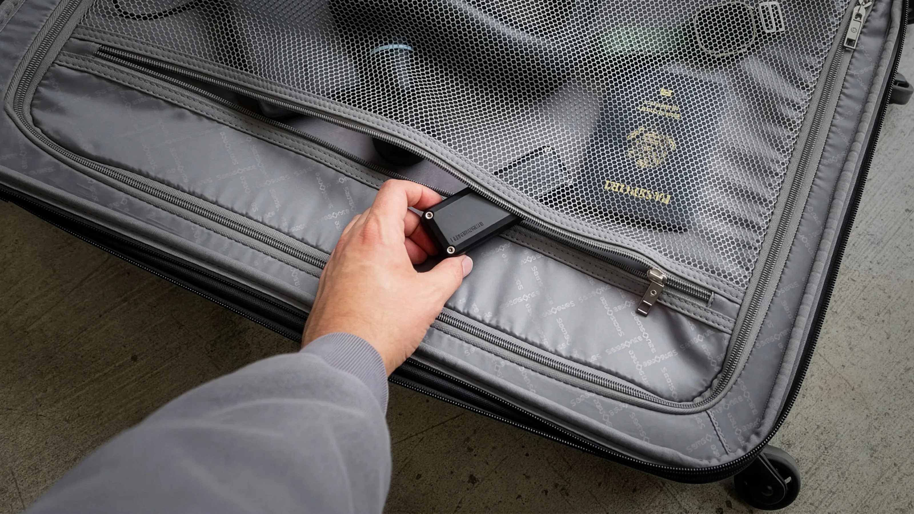 Hand placing a small black device inside a gray suitcase's mesh zippered compartment containing a passport and other items.