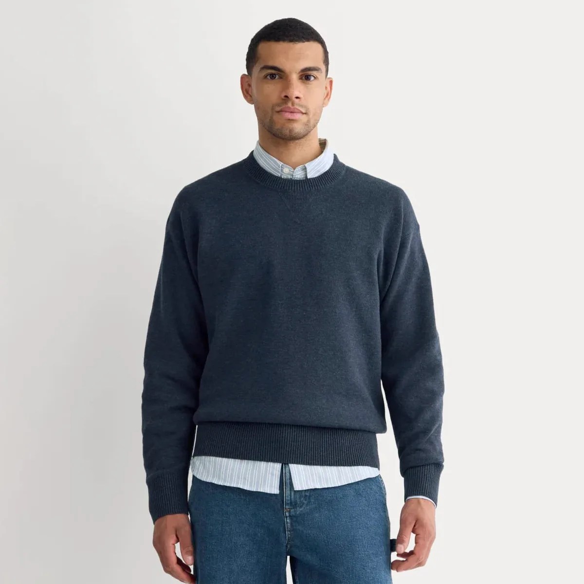 Man wearing a dark blue crewneck sweater over a light blue striped button-up shirt and blue jeans.