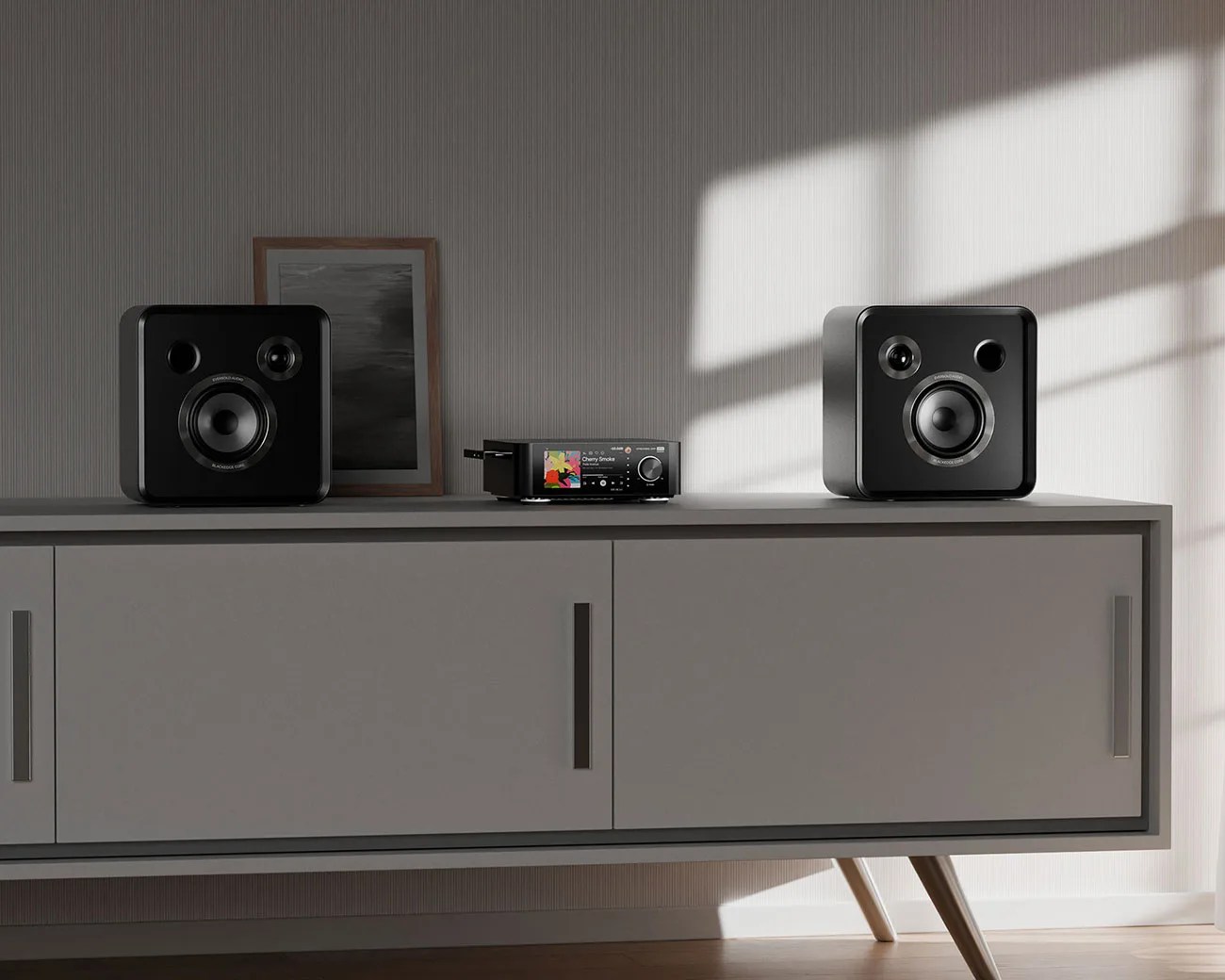 Two black square speakers and a compact stereo receiver with a colorful display on a gray mid-century modern cabinet in a sunlit room.