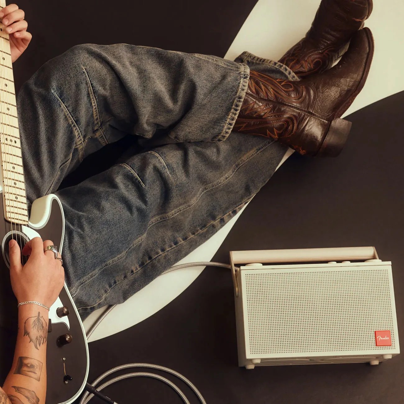 Person wearing brown leather cowboy boots and gray jeans playing a black and white electric guitar next to a beige Fender amplifier.