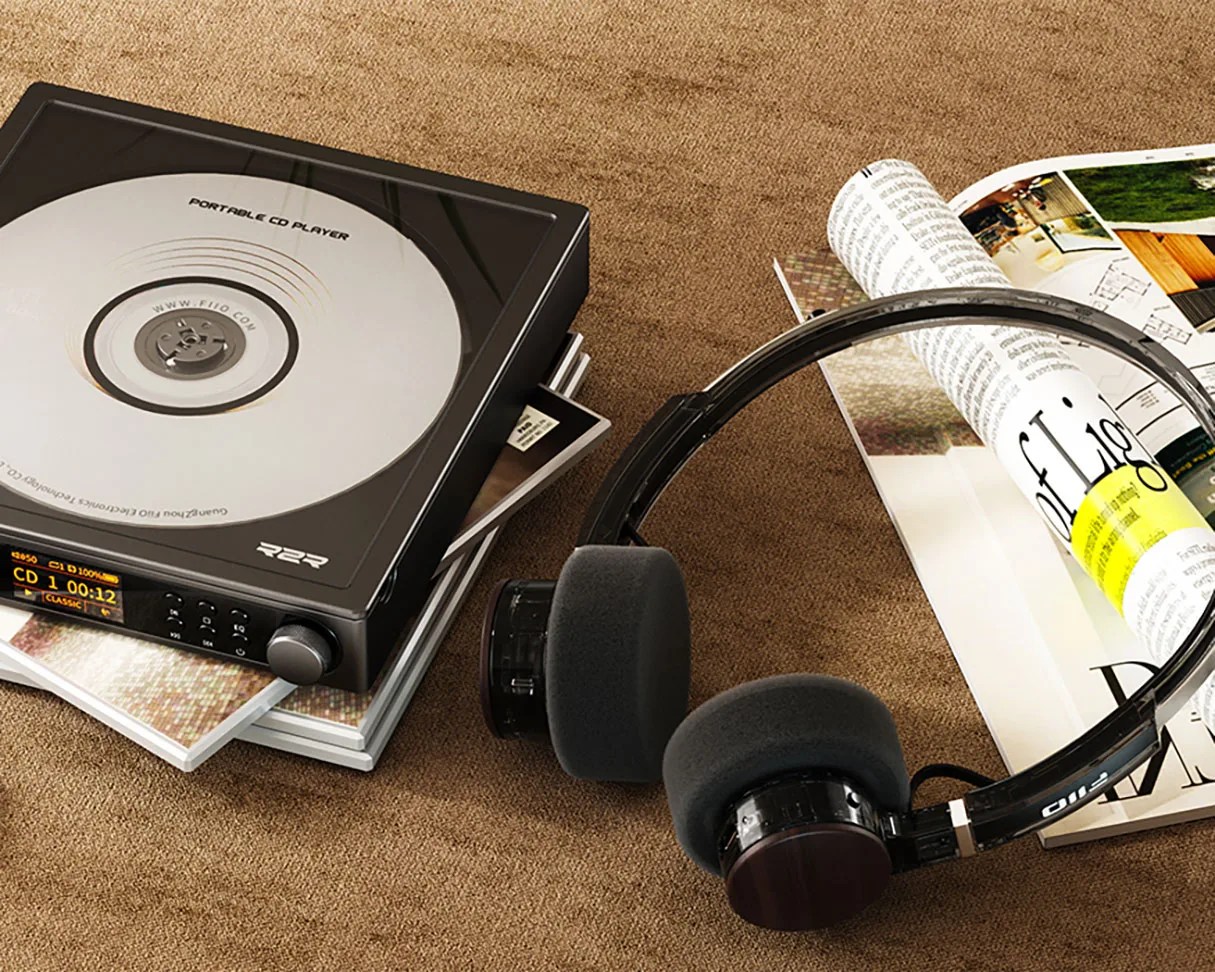 Black portable CD player with a silver disc inside, stacked on CDs next to black on-ear headphones and magazines on a brown surface.
