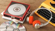Red portable CD player with a clear top, orange foam headphones, and a yellow Silverette typewriter on a brown surface.