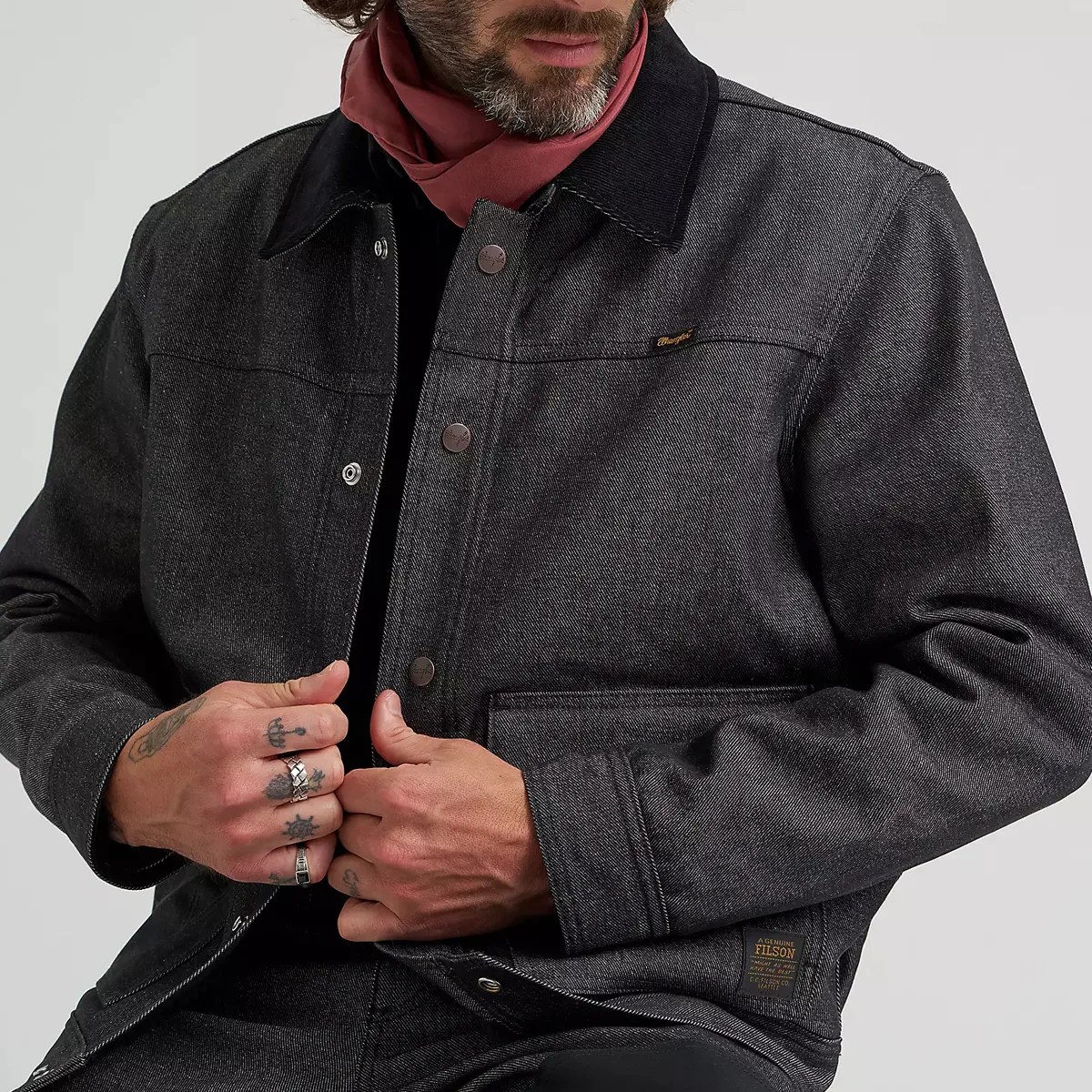 Dark gray denim jacket with black collar and metal snap buttons worn by a person with rings and tattoos on their hands.