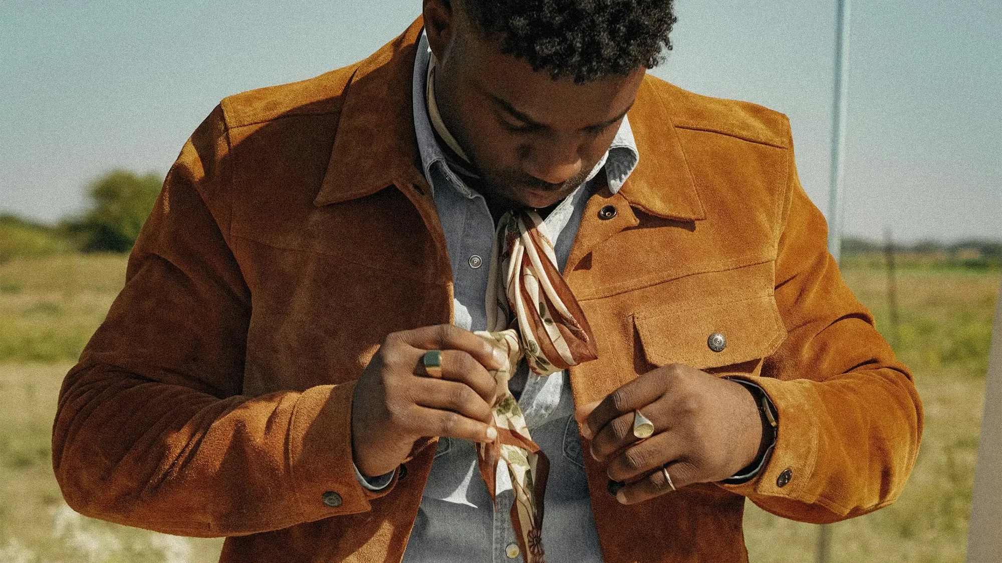 Man wearing a brown suede jacket over a light blue shirt and tying a patterned scarf around his neck.