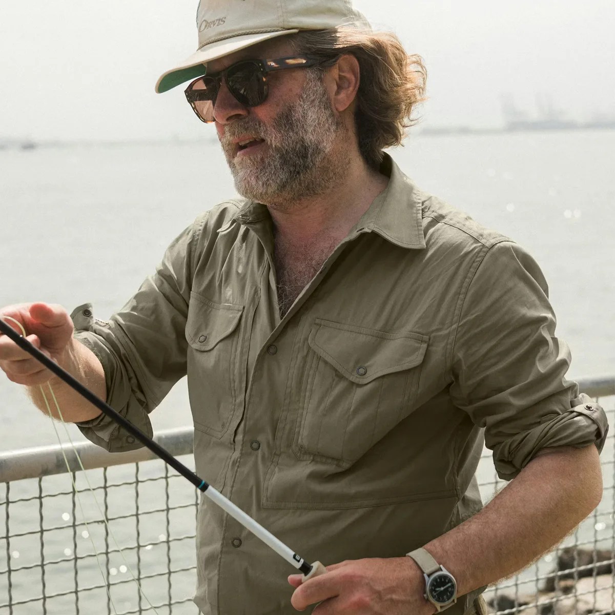 Man wearing a beige fishing shirt and cap, holding a fishing rod near a waterfront railing.
