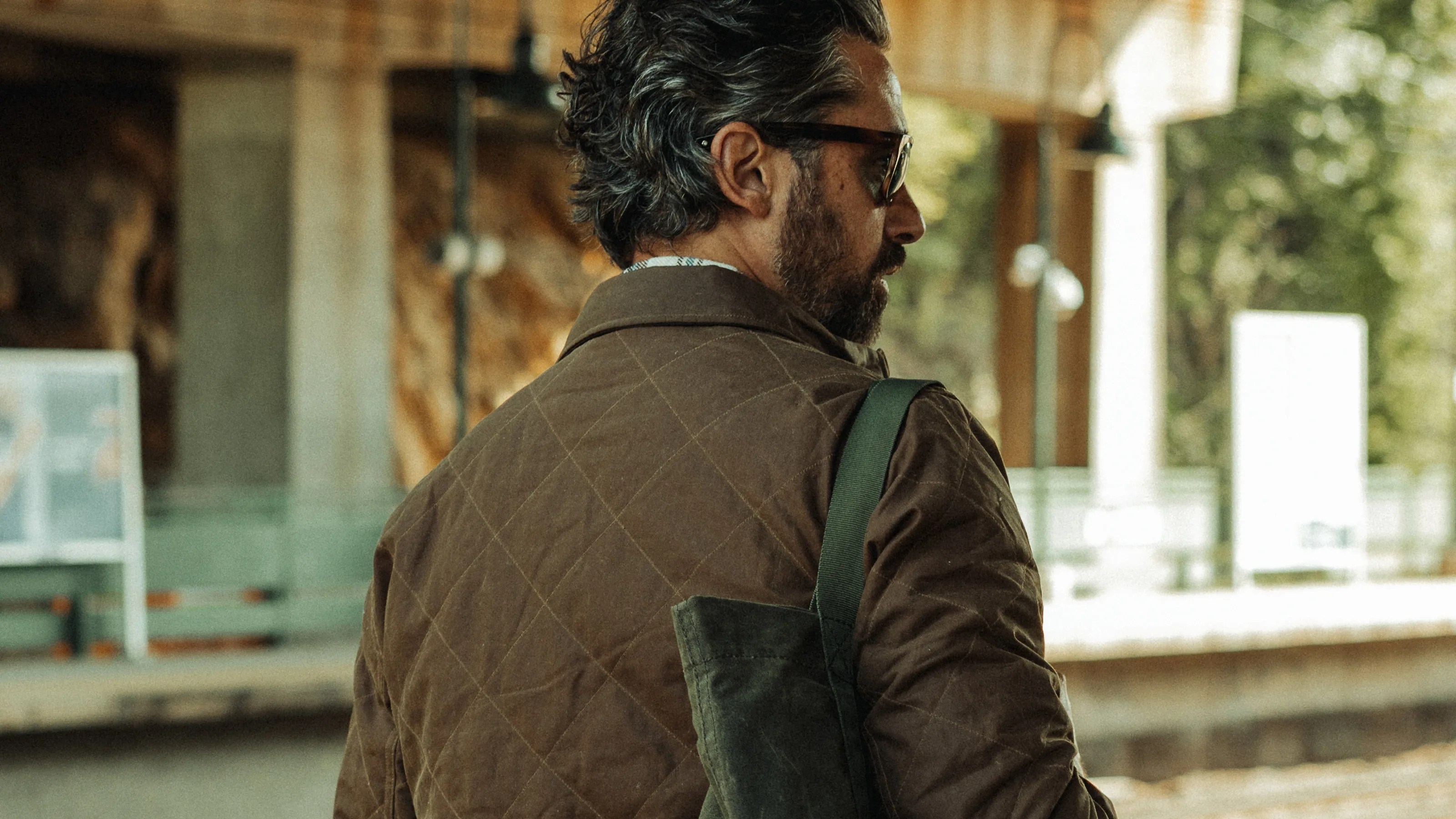Man with dark hair and beard wearing a brown quilted jacket and carrying a green shoulder bag.