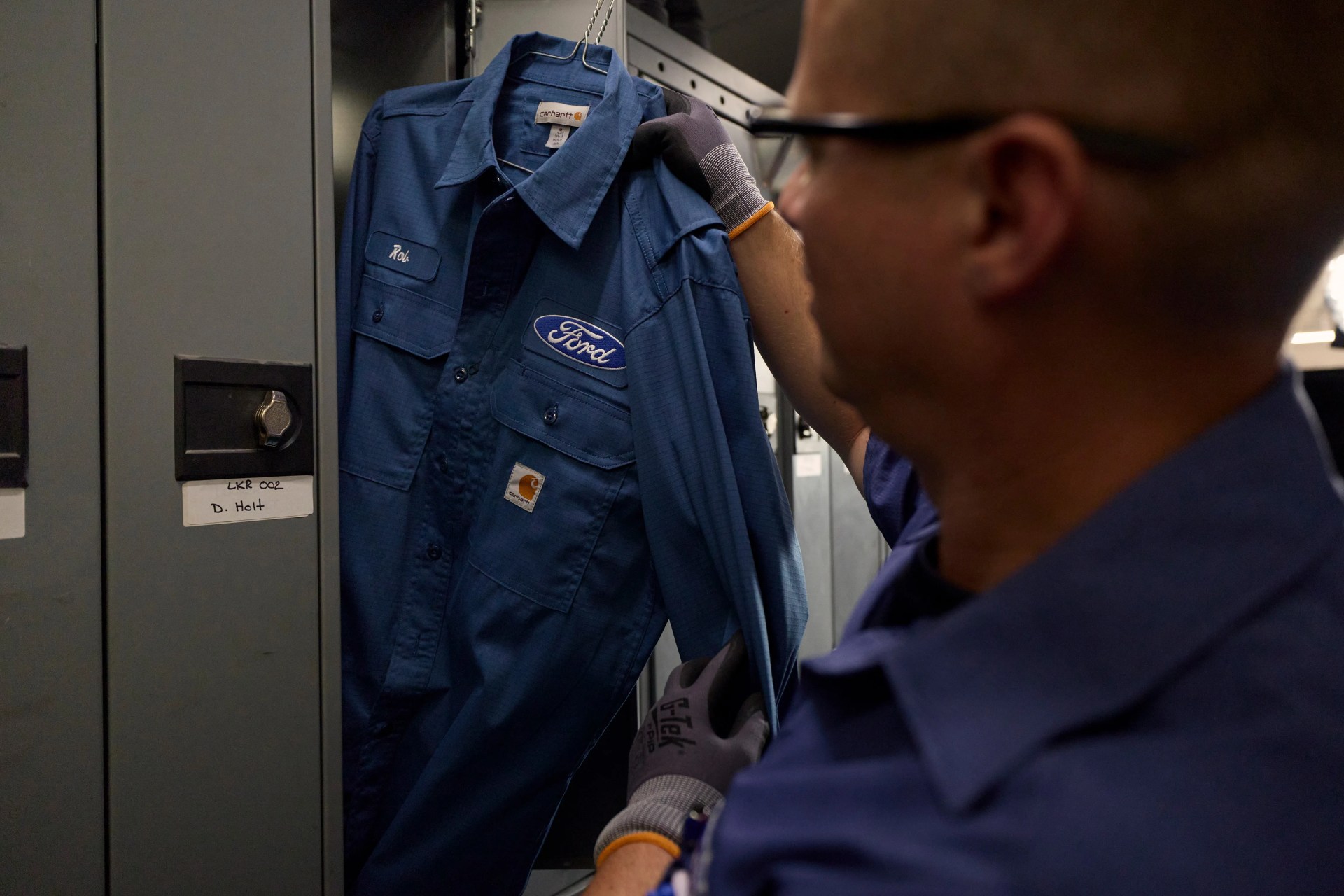 Person wearing gloves holding a blue Carhartt work shirt with Ford logo on a hanger near lockers.