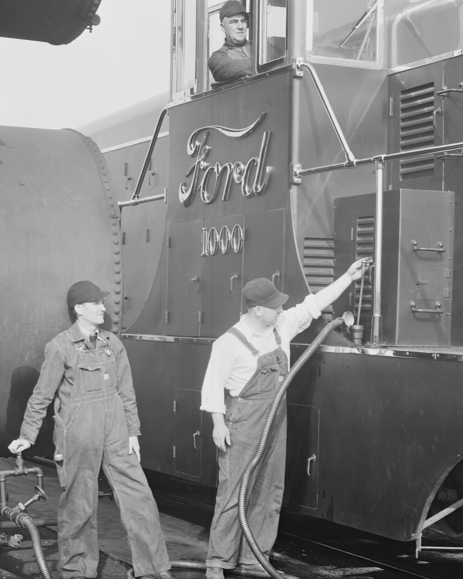 Three men in work overalls and caps servicing a large Ford 1000 industrial machine with hoses attached.