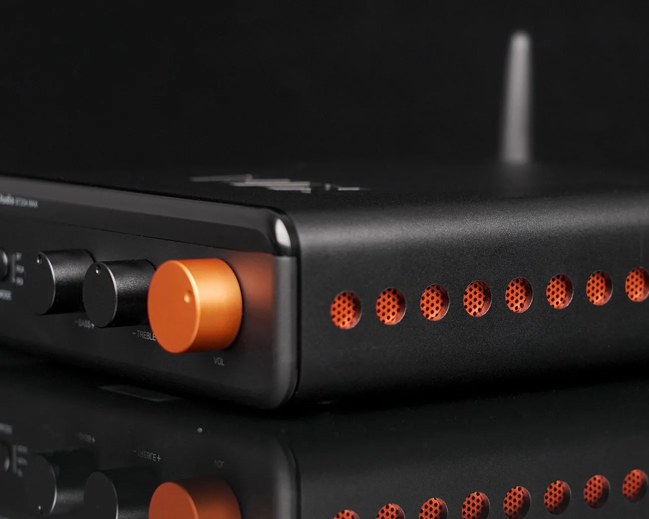 Black audio amplifier with orange volume knob and orange perforated side vents on a reflective surface.