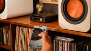 Hand holding a smartphone with a music app, pointed at a black audio amplifier with orange knob on a wooden shelf between white speakers.