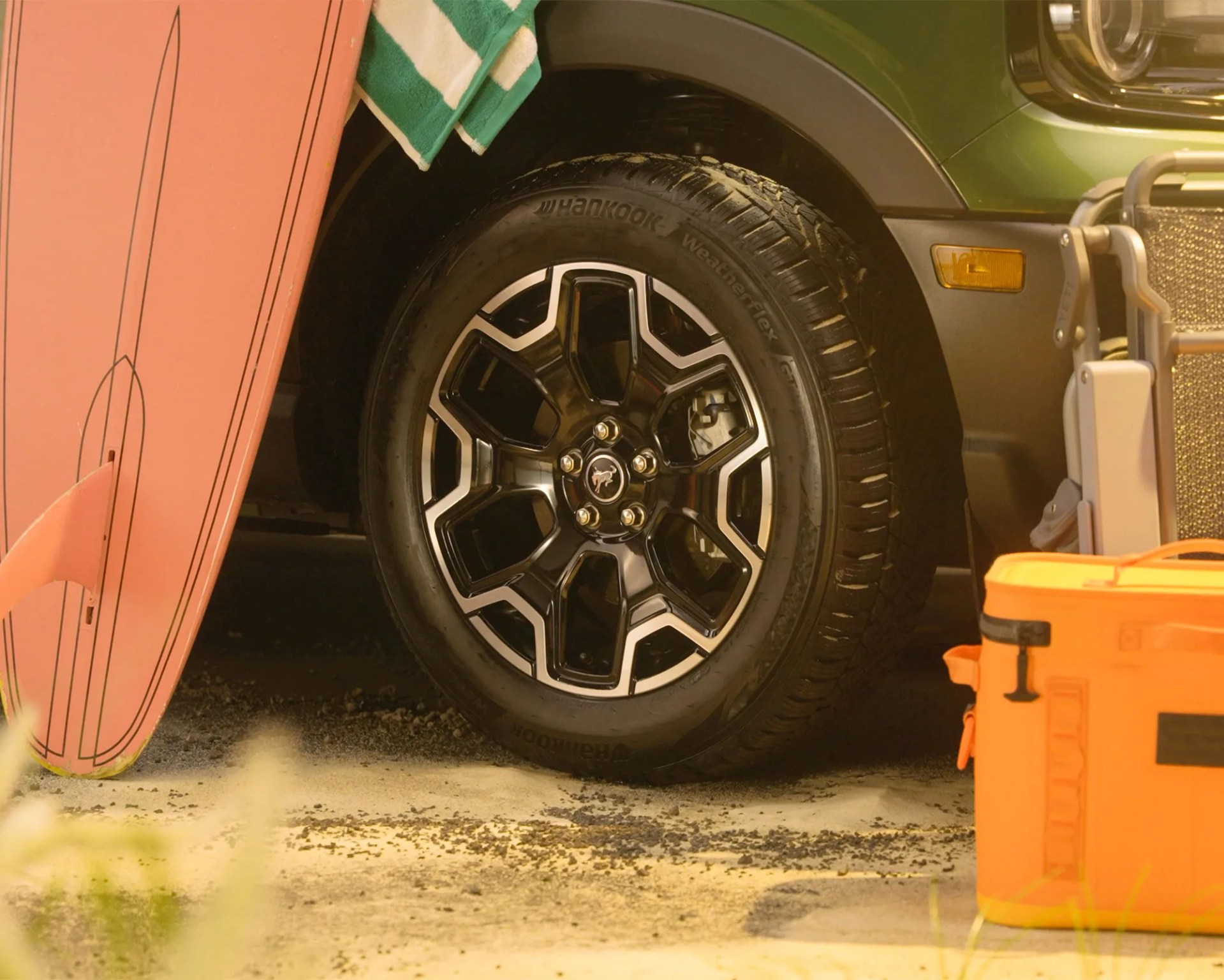 Black and silver alloy wheel with Hankook Weatherflex tire on a green vehicle next to a pink surfboard and orange cooler.