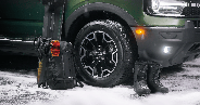 Green off-road vehicle front with black tire, black backpack with orange goggles, black gloves, skis, and black snow boots on snow.