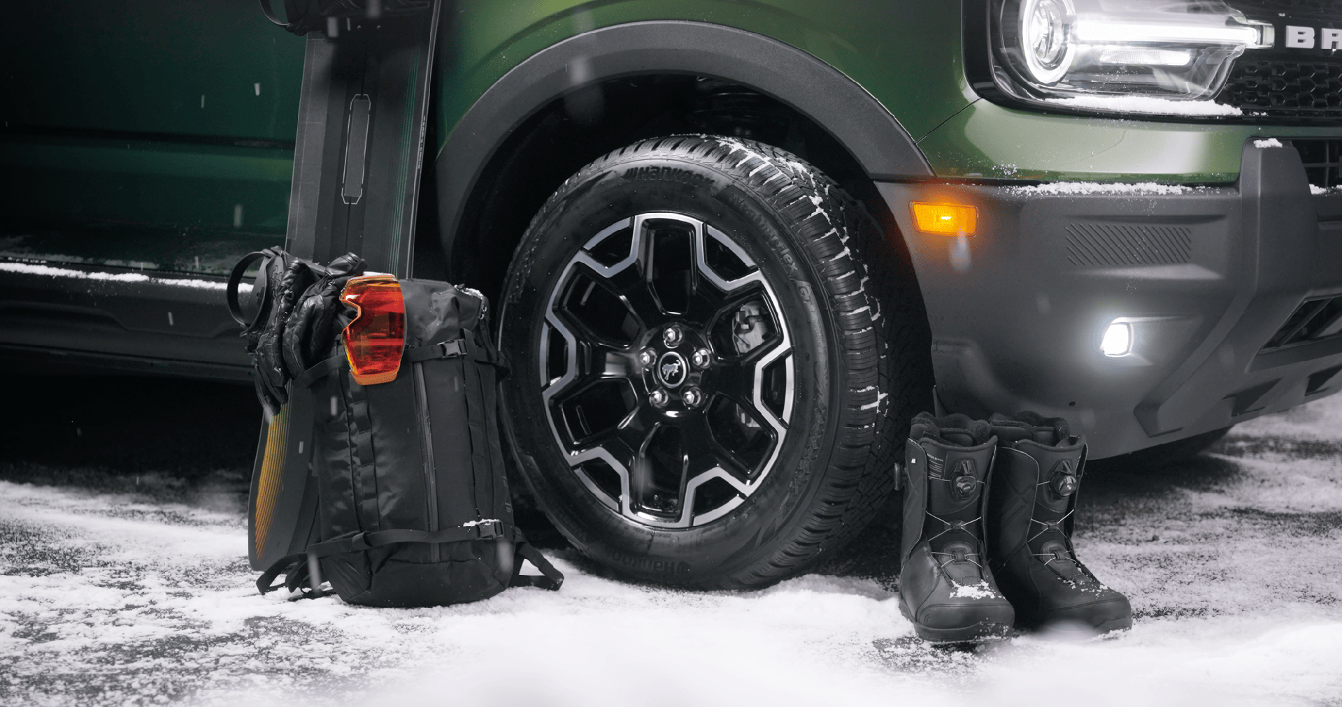 Green off-road vehicle front with black tire, black backpack with orange goggles, black gloves, skis, and black snow boots on snow.