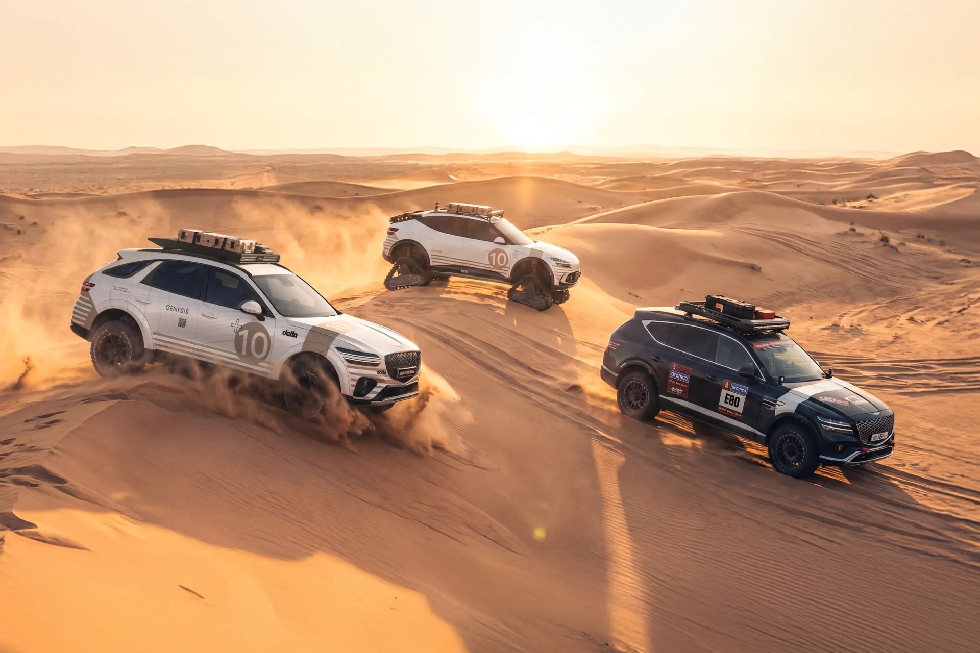 Three off-road SUVs, two white and one dark blue, driving on sand dunes at sunset, with one SUV equipped with tank-like tracks.