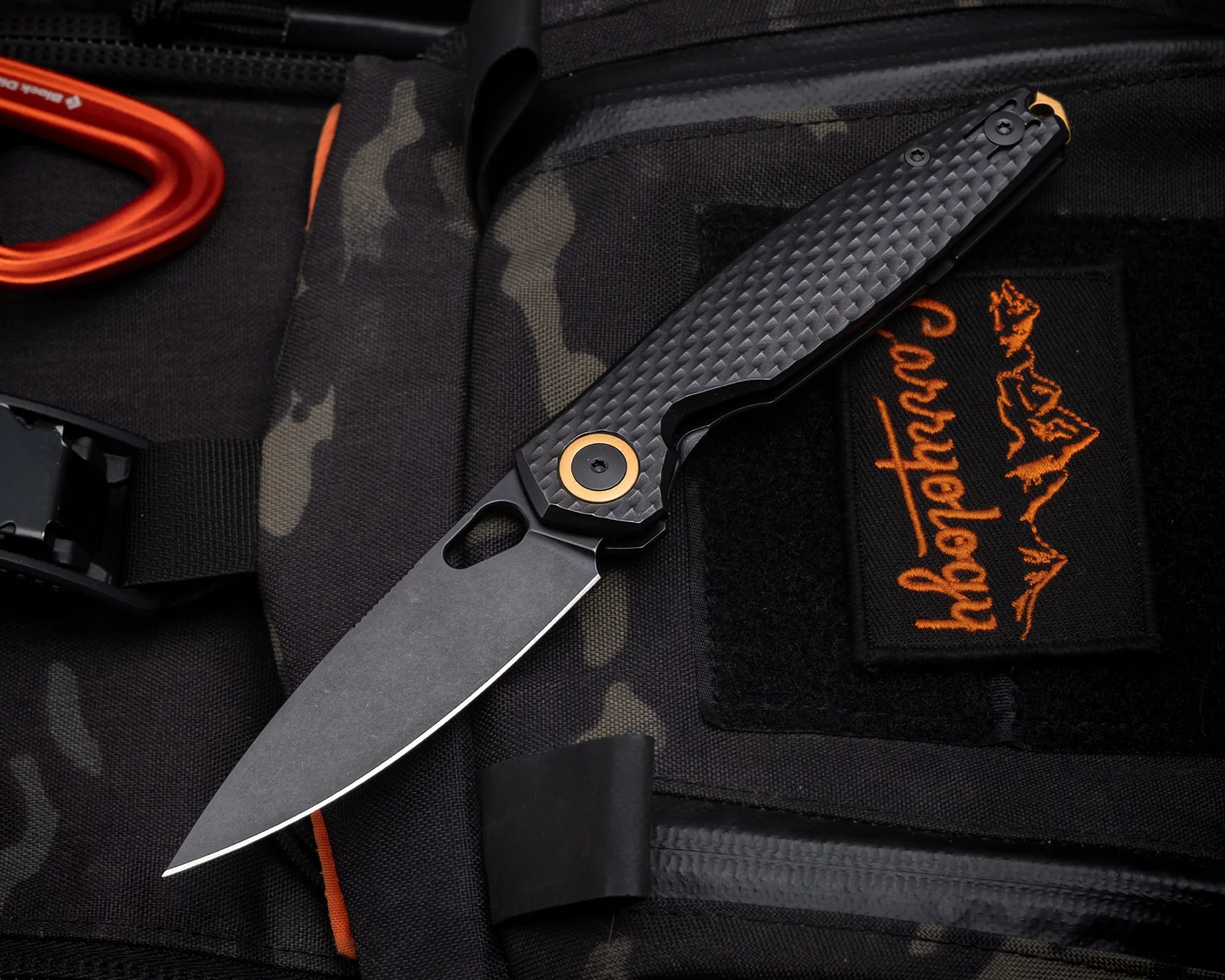 Black folding knife with textured handle and gold pivot detail on a black and camo fabric surface.
