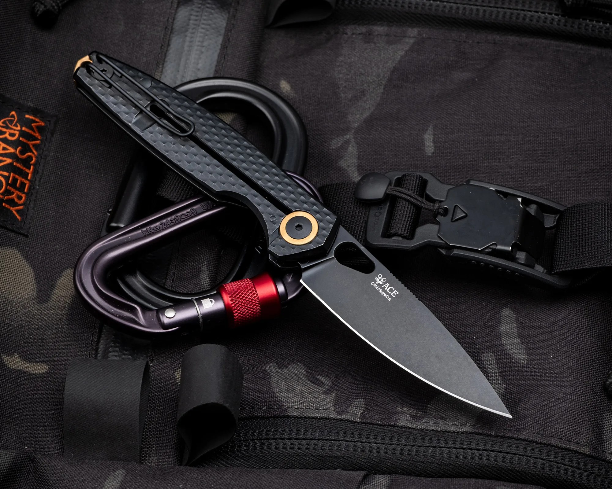 Black folding knife with a textured handle and gold pivot, resting on a black carabiner and dark fabric with "Mystery Ranch" label.