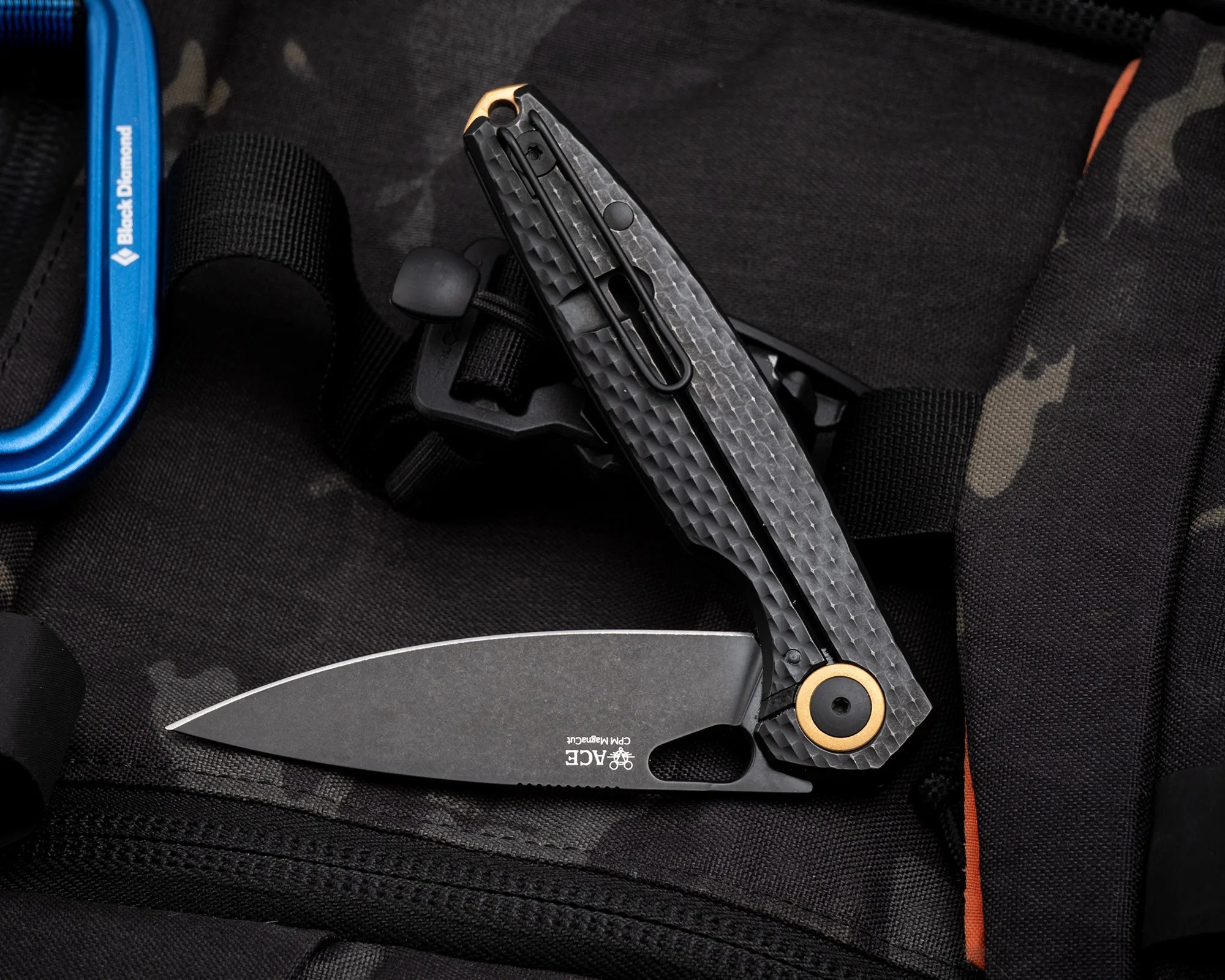 Black folding knife with textured handle and gold pivot, partially open on a black fabric surface with a blue carabiner nearby.