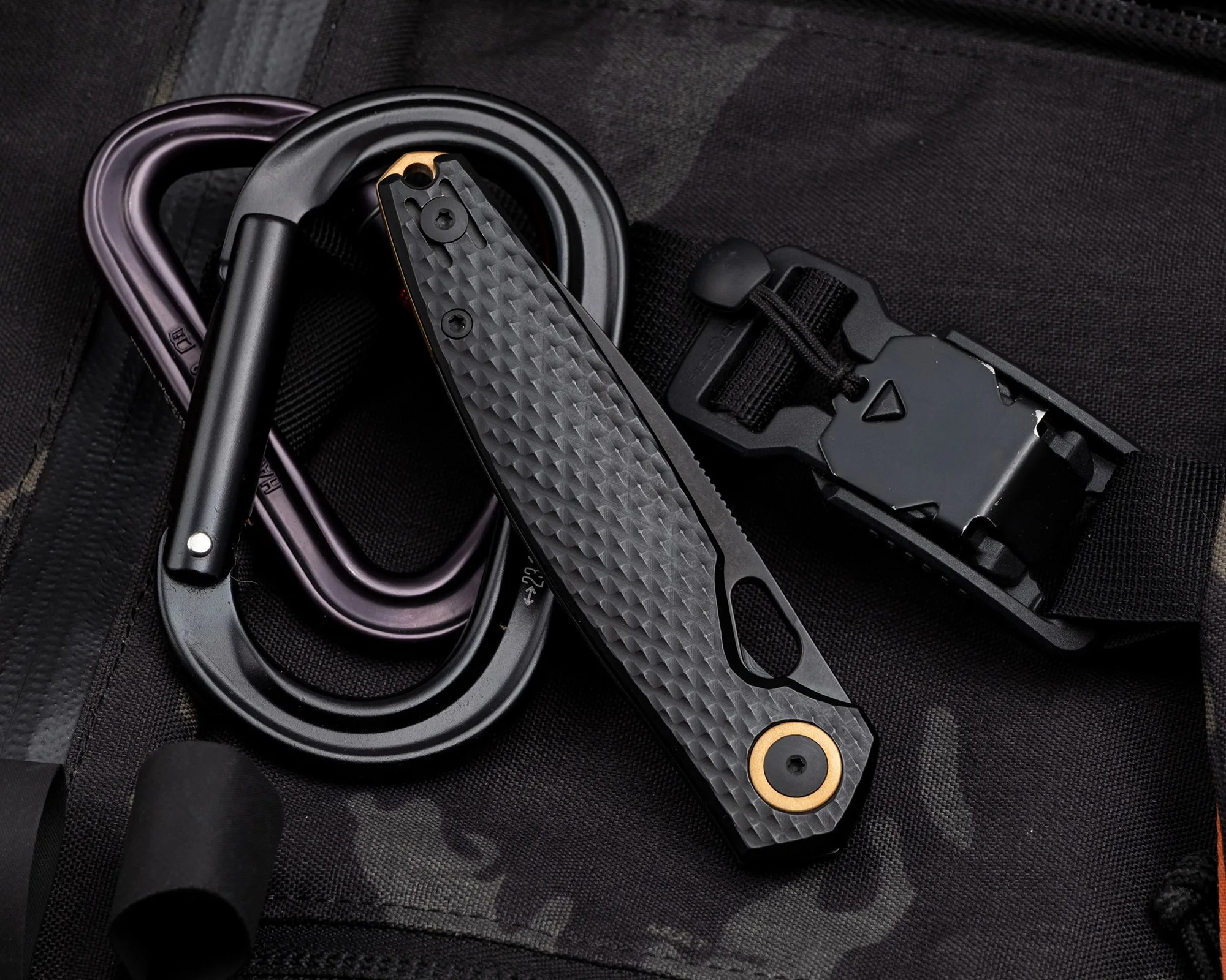 Black textured folding knife with bronze pivot and two black carabiners on black fabric.
