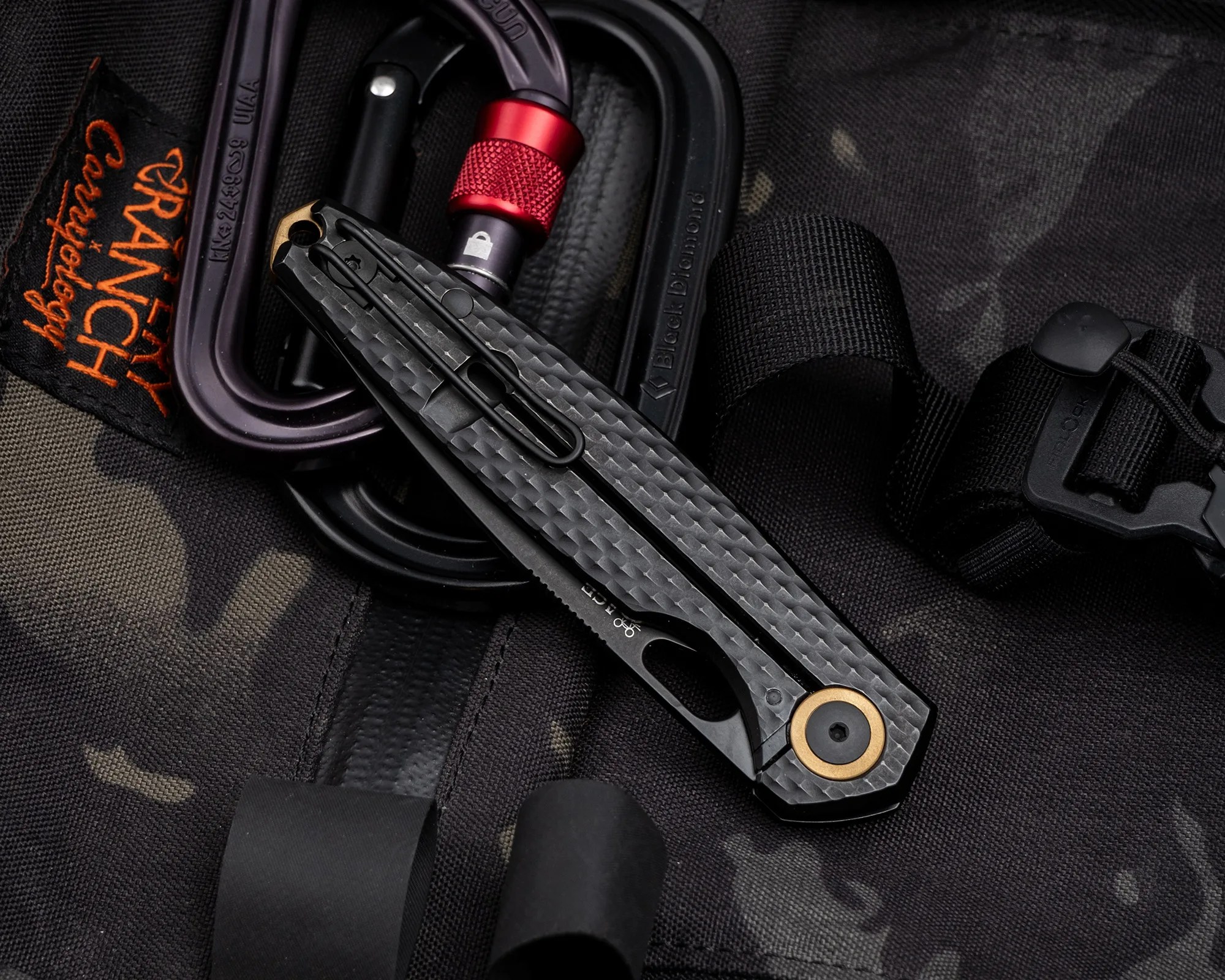 Folded black textured pocket knife with a brass pivot on a camo fabric next to black and purple carabiners.